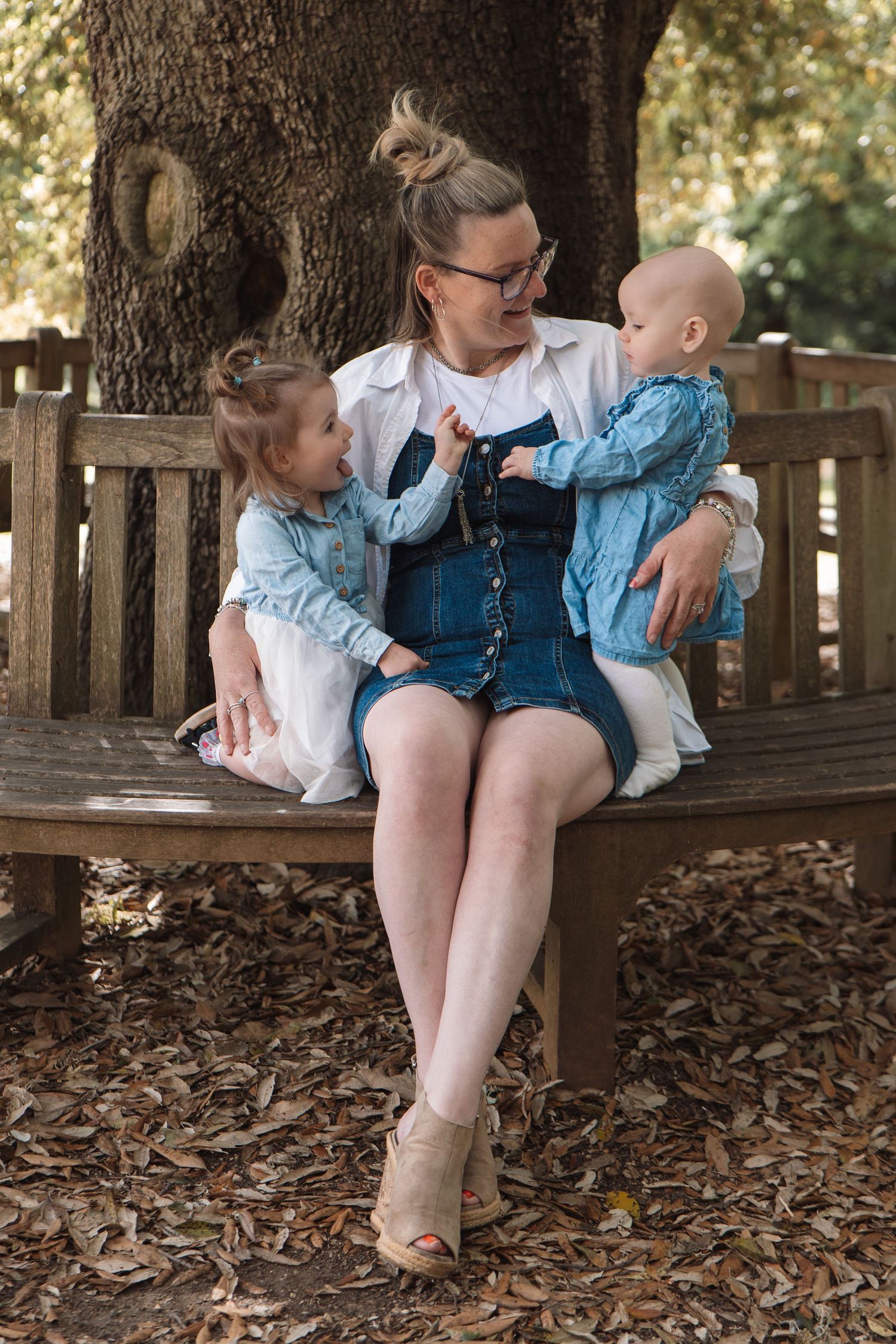 A woman sits on a wooden bench with two young children in a park, all wearing denim outfits among autumn leaves.