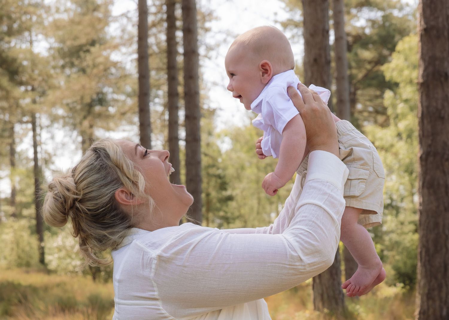 A joyful mother lifts her smiling baby in the air outdoors surrounded by tall pine trees.