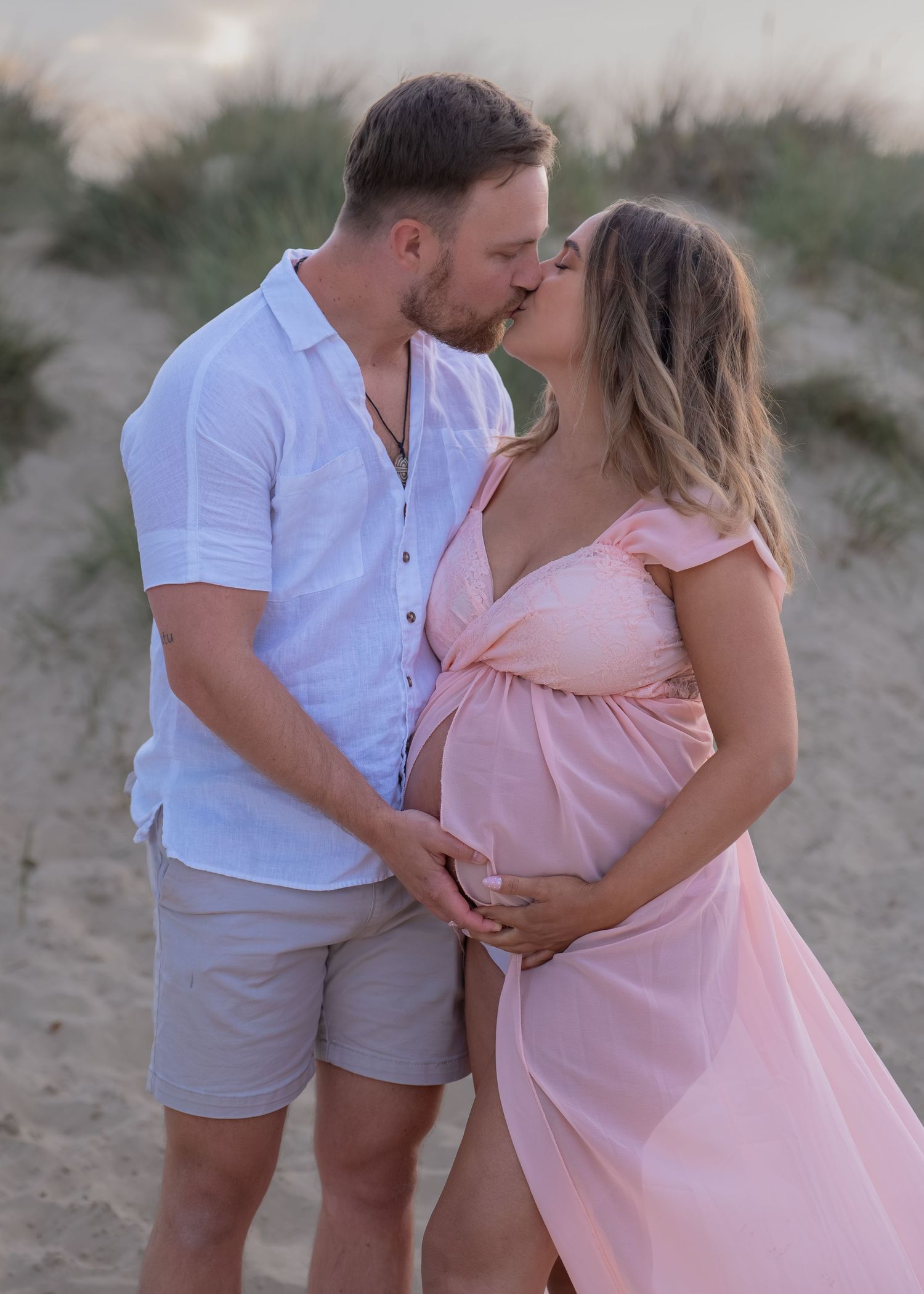 Couple sharing a kiss on sandy beach dunes, pregnant woman in pink maternity dress held by man in white shirt.
