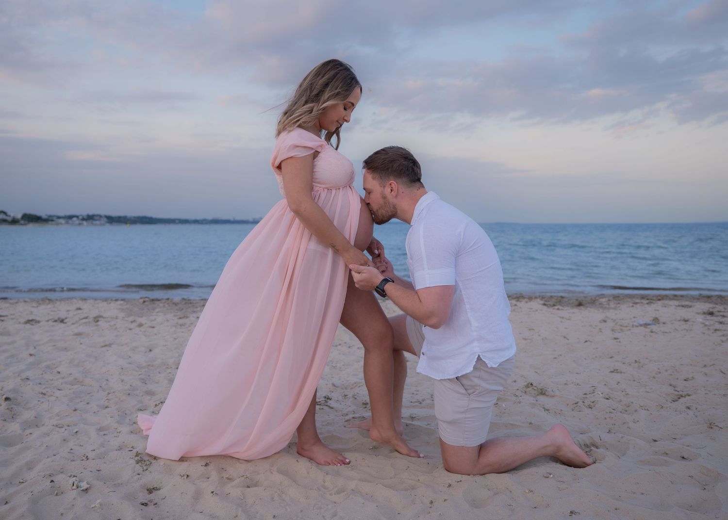A man kneeling on a sandy beach kissing a pregnant woman's belly in a pink maternity gown at sunset.