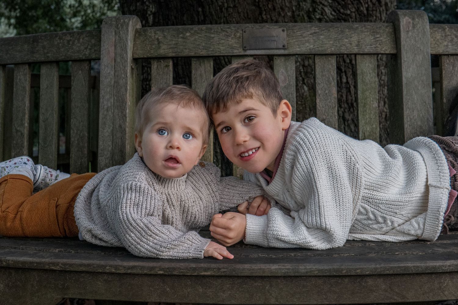 Two young brothers in matching knit sweaters lying together on a wooden park bench, smiling at the camera.