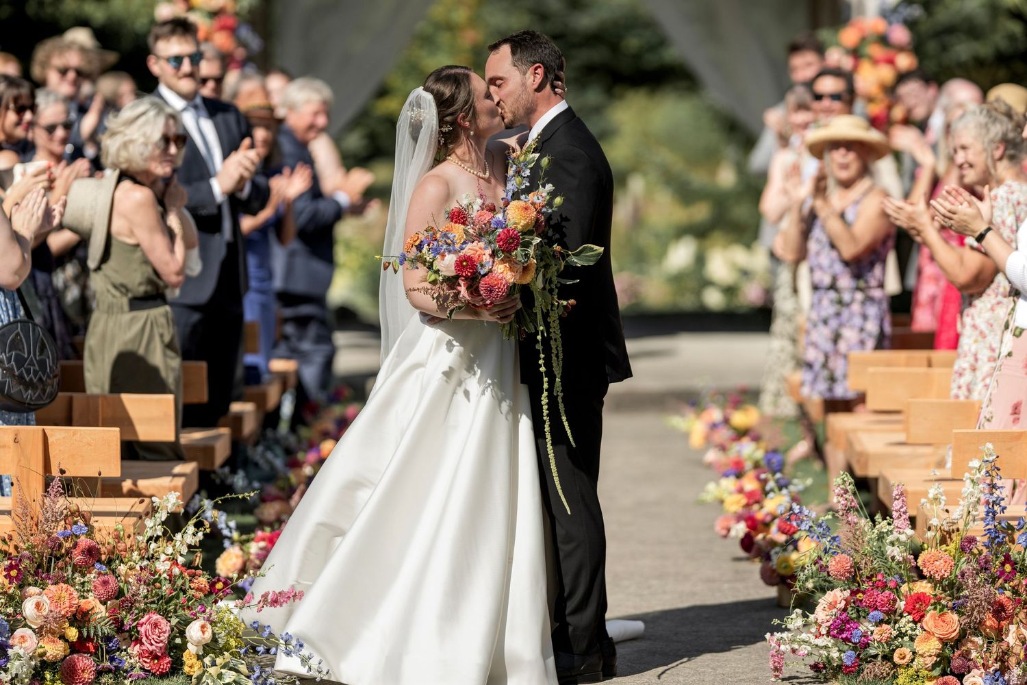 Bride and groom sharing first kiss at colorful floral-lined wedding ceremony aisle outdoors.
