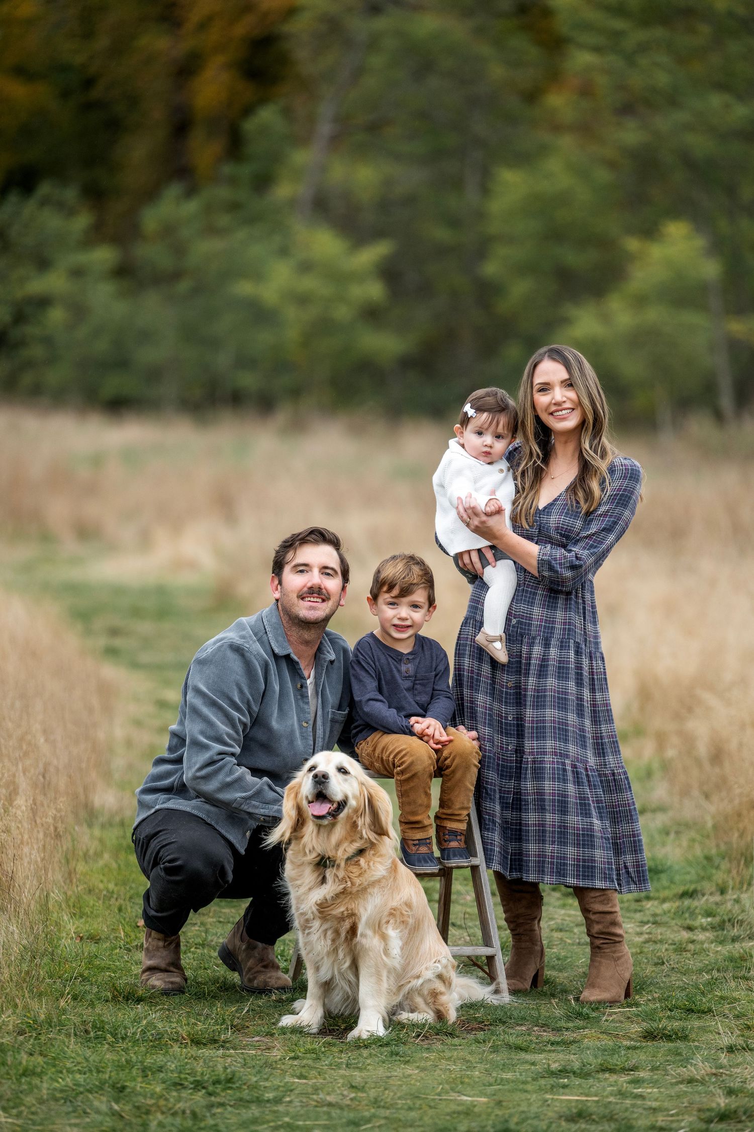 Happy family with golden retriever poses in autumn field, parents with two young children in casual outdoor portrait.