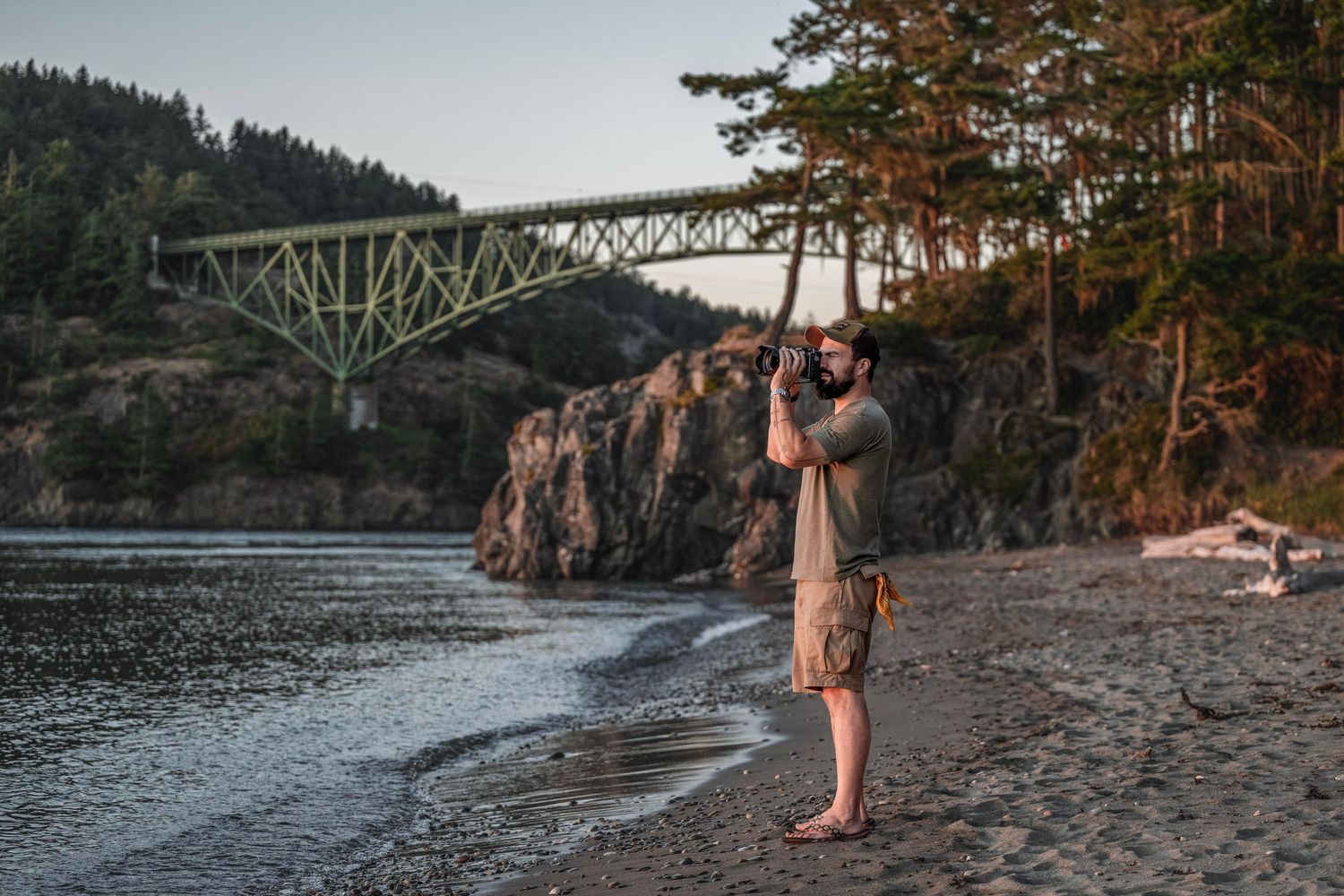 Man photographing scenic coastal landscape with green bridge and rocky cliffs at golden hour on a pebble beach.