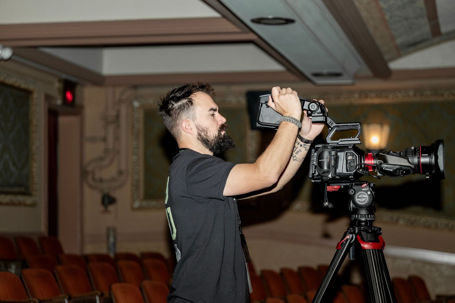 Bearded videographer operating professional camera on tripod inside ornate theater with red seats.
