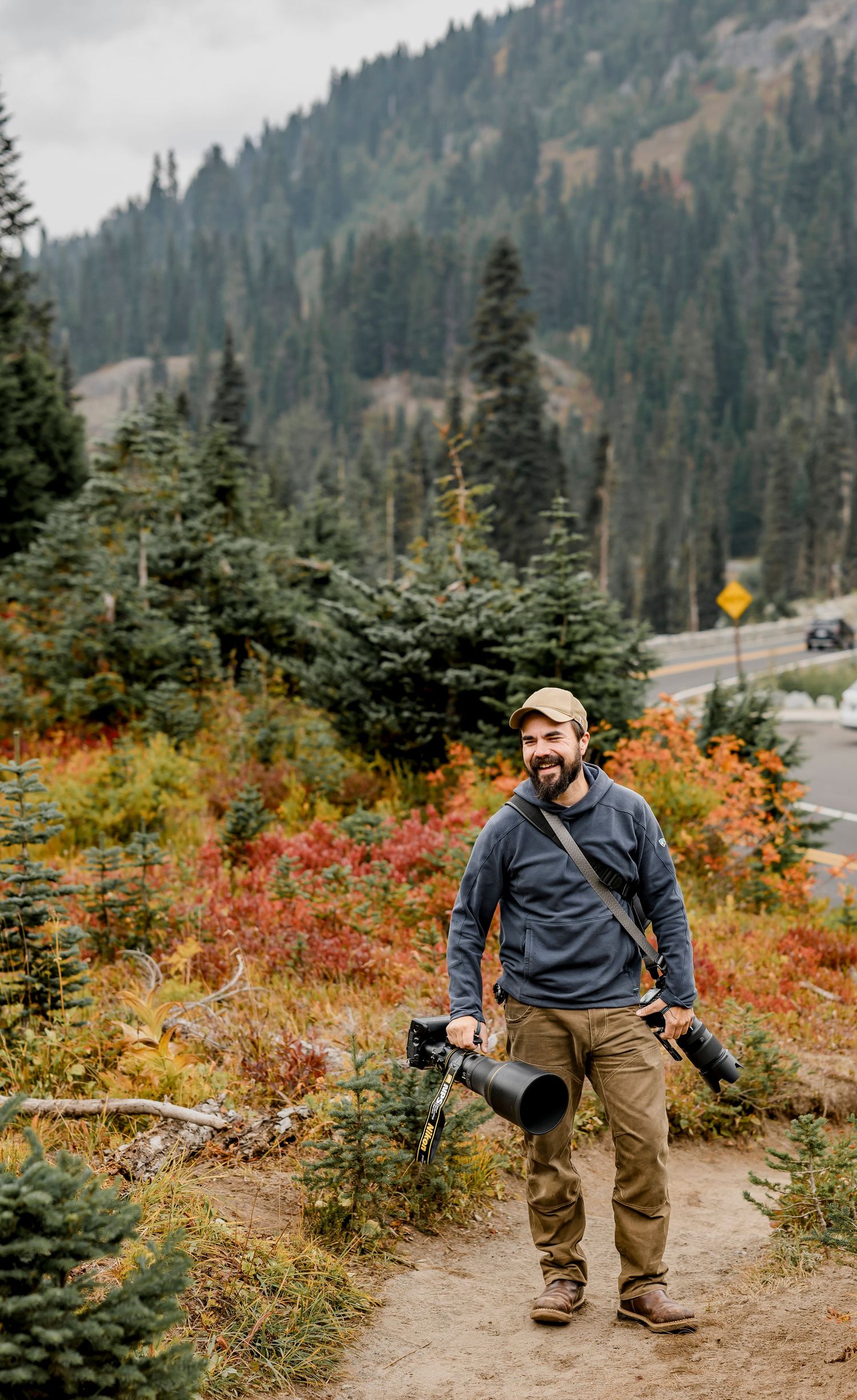 Smiling bearded male photographer with cameras hikes trail amid vibrant fall foliage and pine forest.