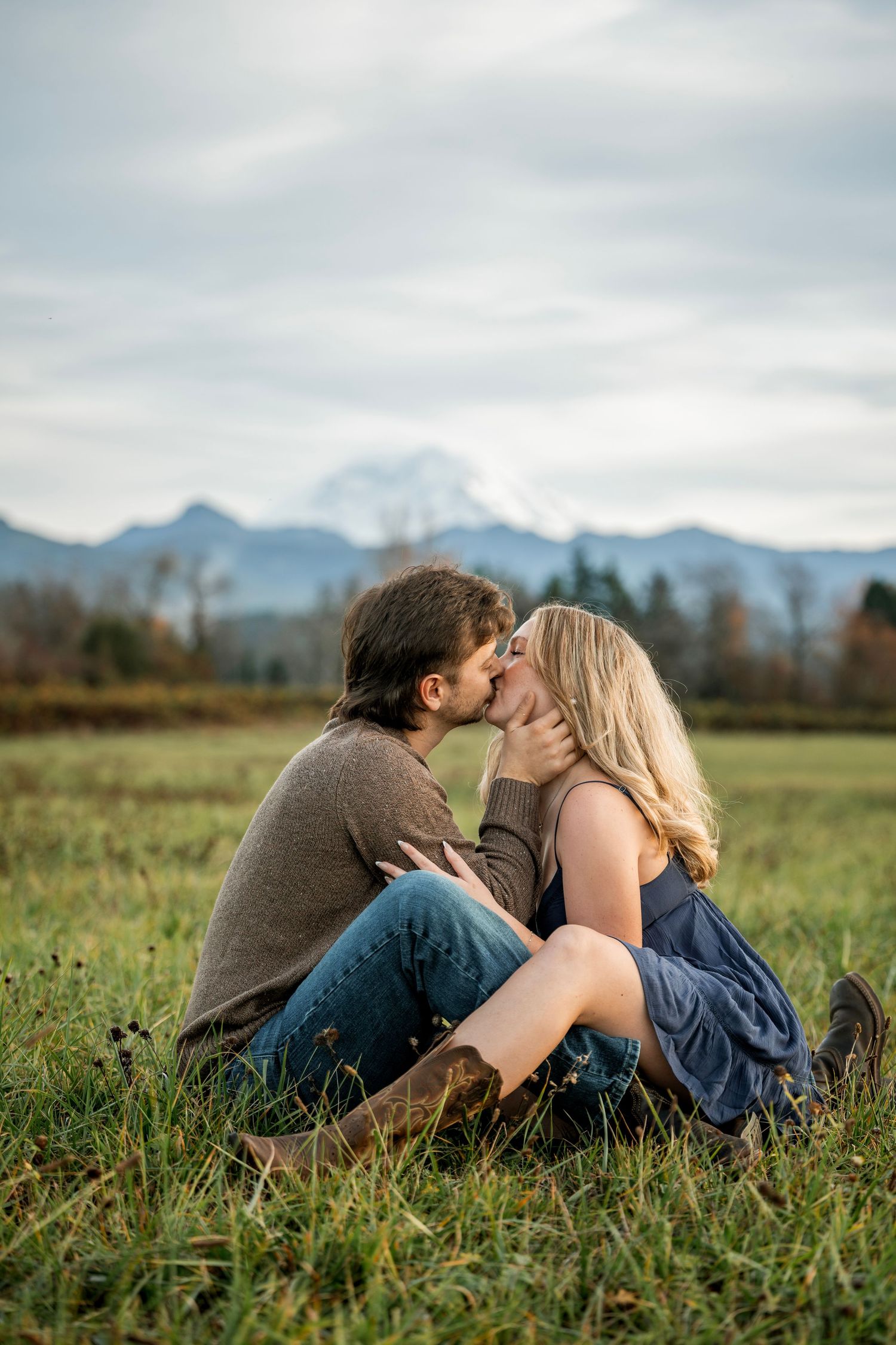 Couple kissing while sitting in green field with mountain backdrop on cloudy day.