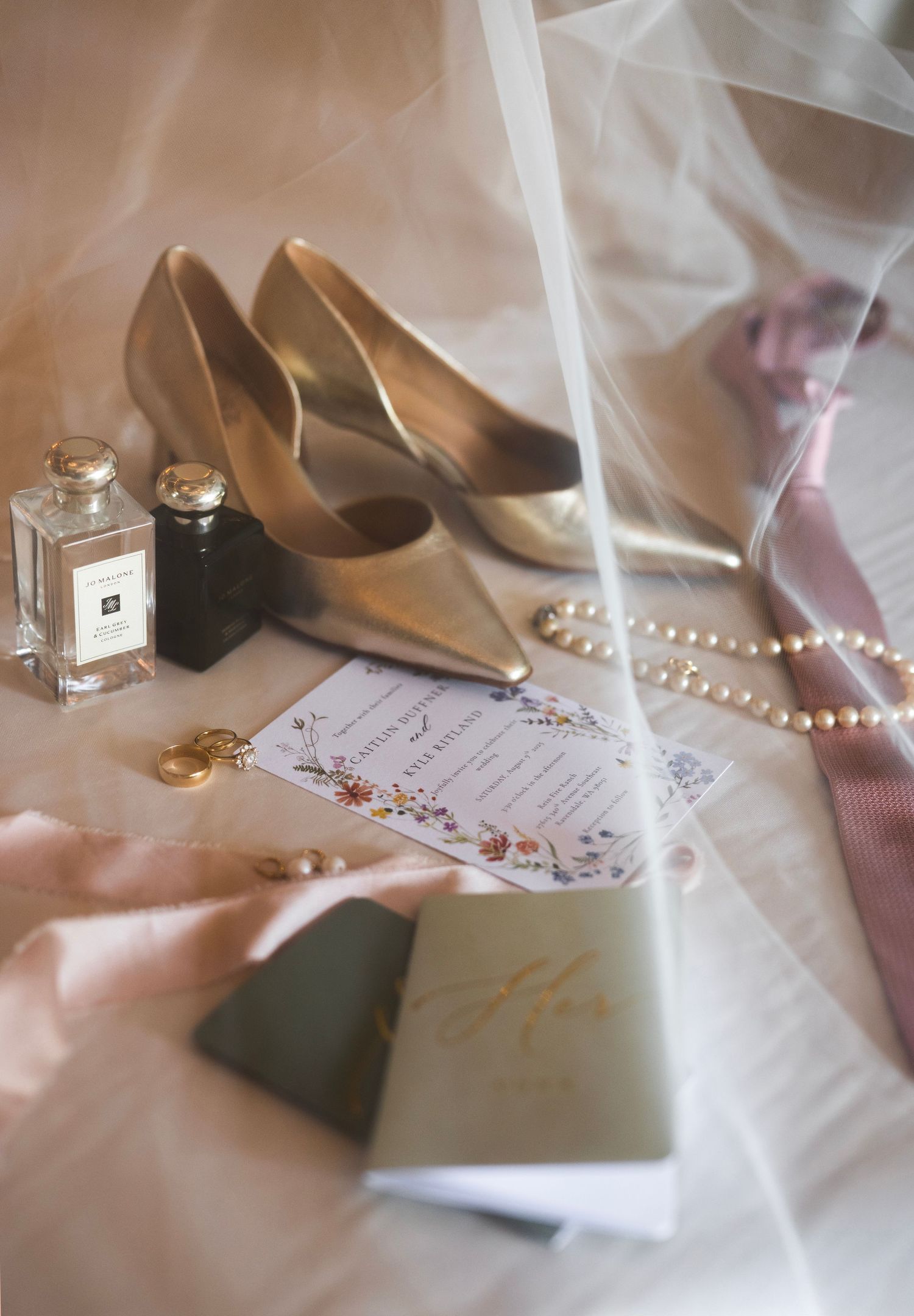 Gold bridal heels, Jo Malone perfume, pearl necklace, floral wedding invitation, and veil arranged on a soft surface.