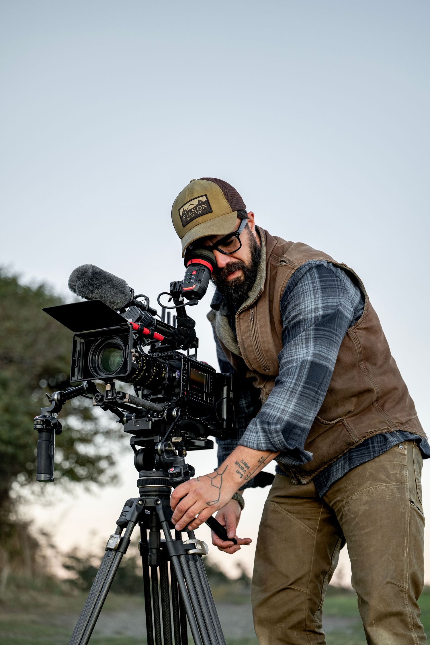 Bearded male filmmaker adjusting a professional cinema camera on a tripod in an outdoor setting.