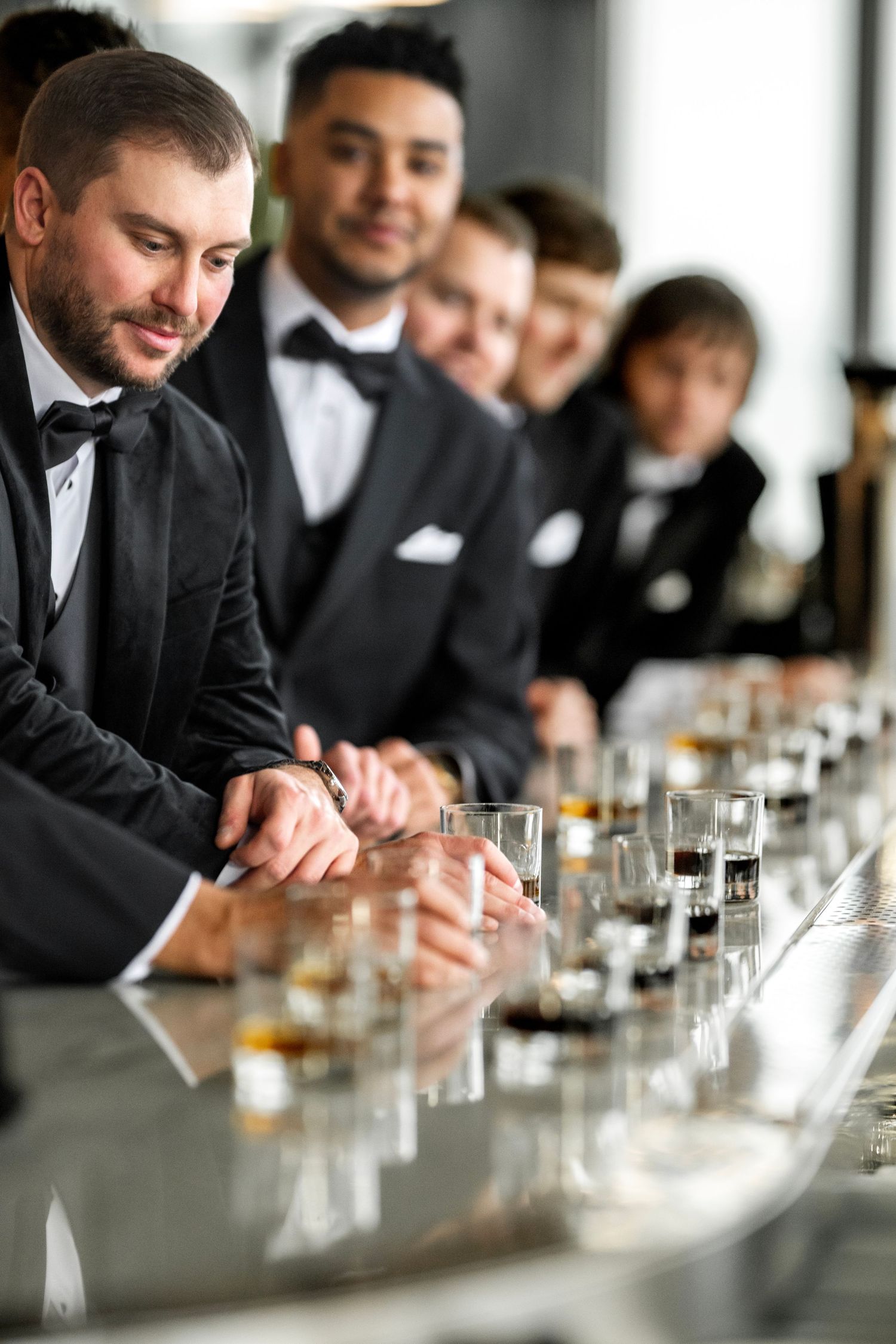Groomsmen in black tuxedos lined up at bar with whiskey shot glasses.