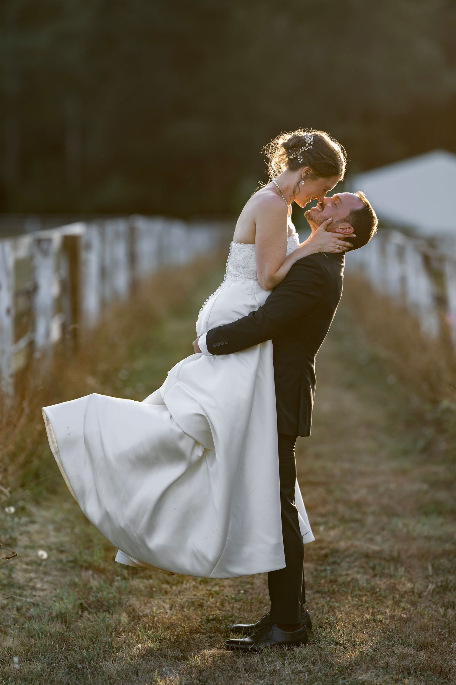 Groom lifting bride in white gown at golden hour near rustic fence outdoors.