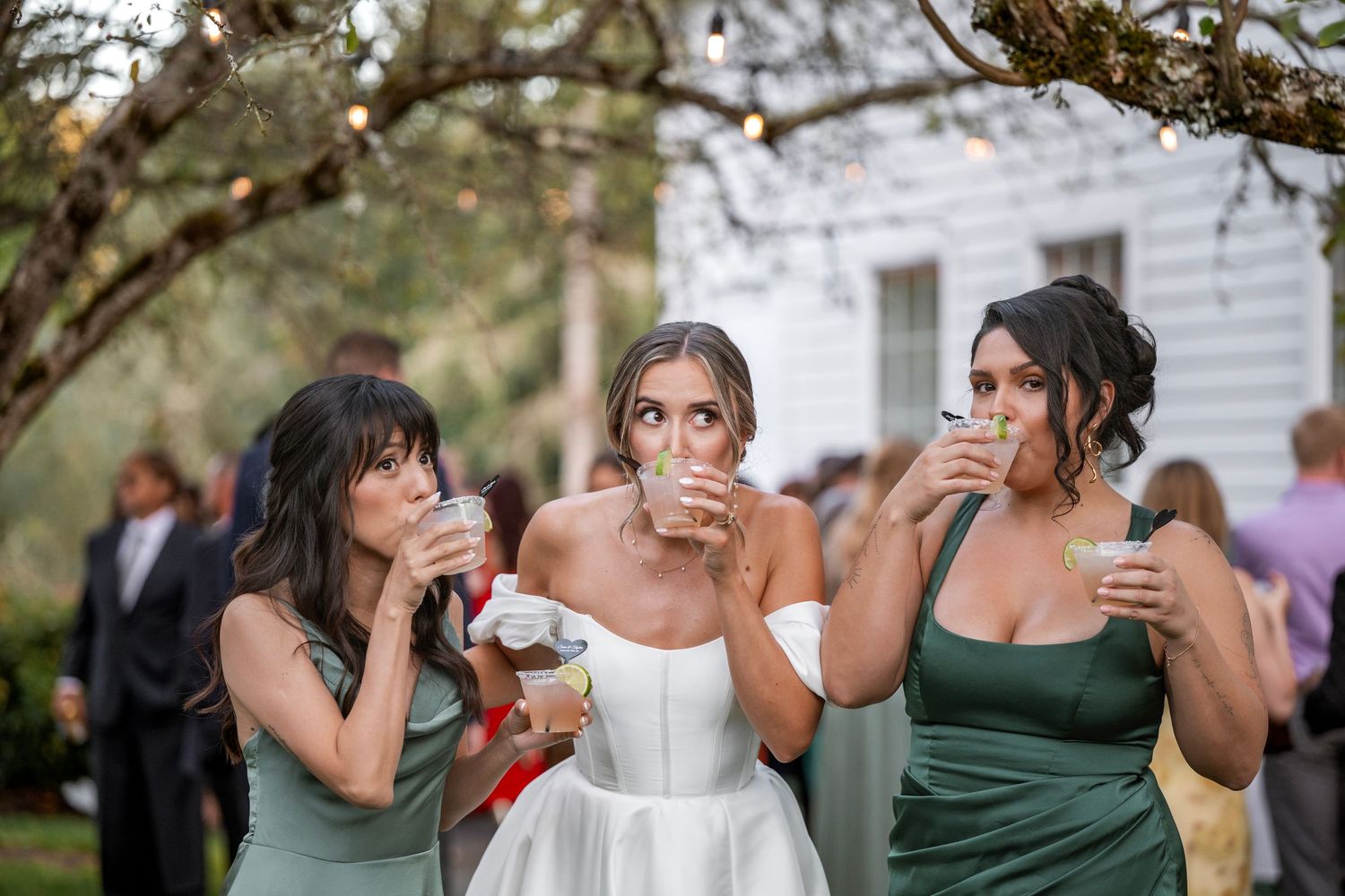 Bride in white gown and bridesmaids in sage green dresses toasting drinks at outdoor wedding reception.