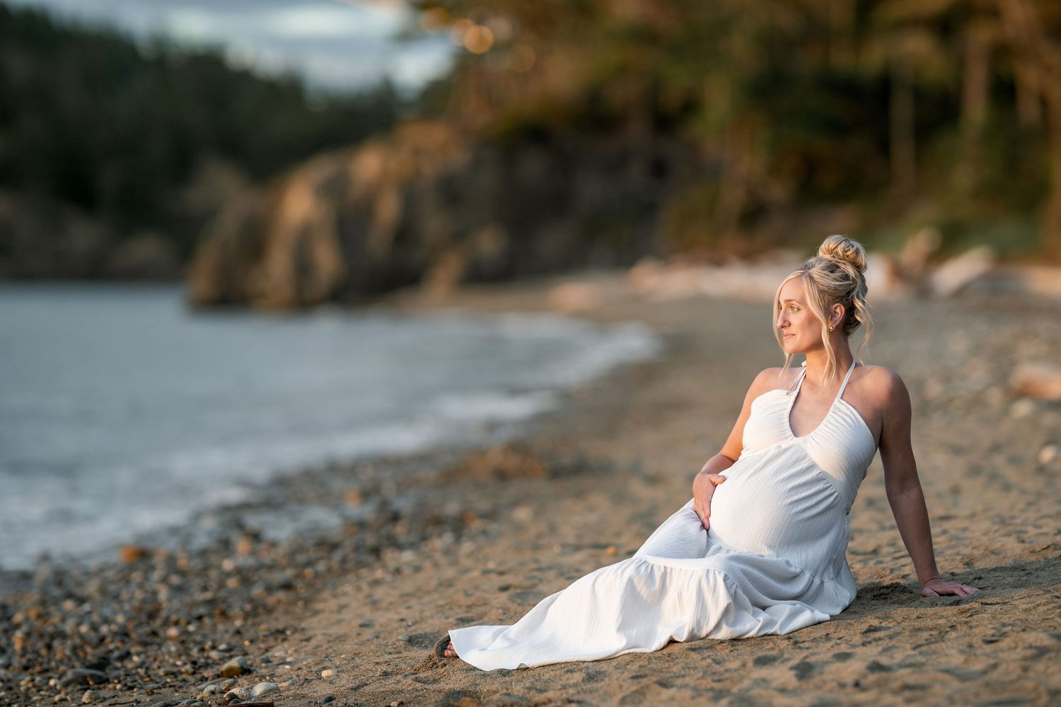 Pregnant woman in white dress sitting peacefully on a rocky beach shore with trees and water in the background.