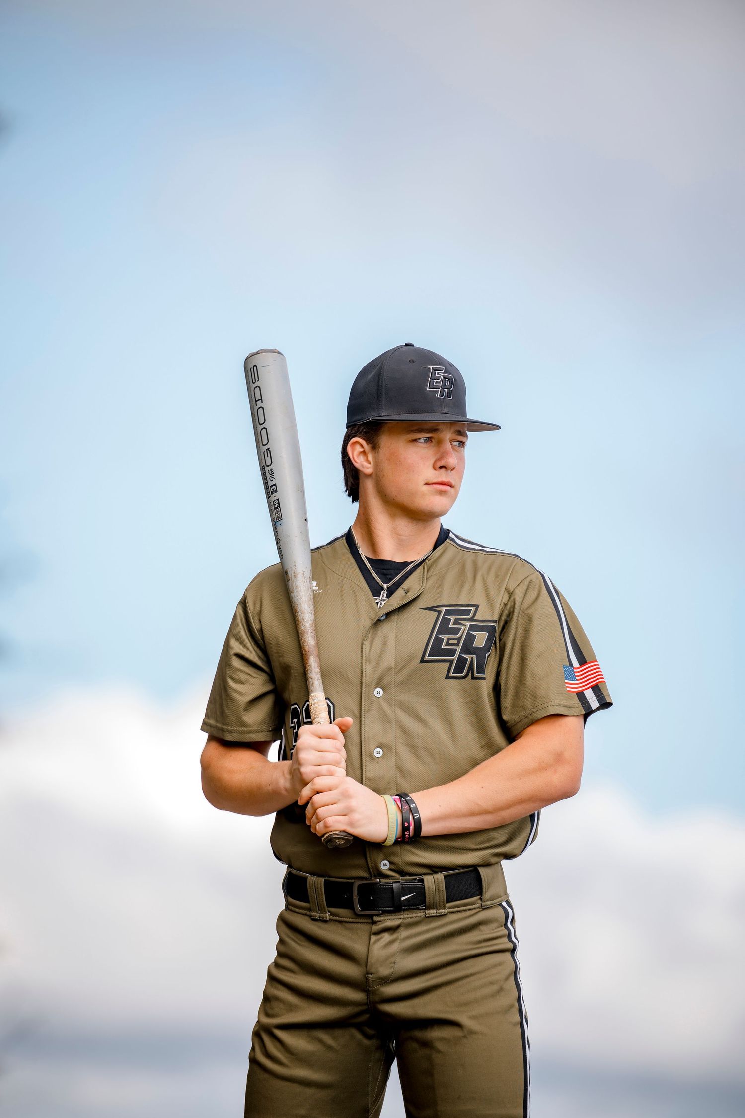 Young baseball player in olive uniform holding a bat against a cloudy sky backdrop.