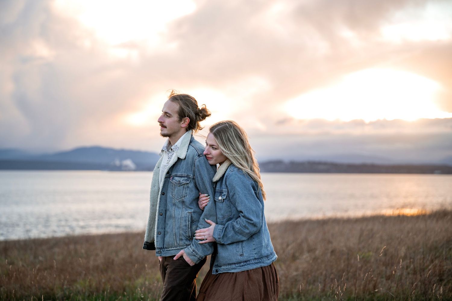 Couple in denim jackets embracing in a field at sunset by the water.