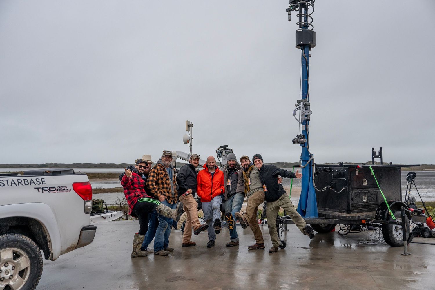 Group of people huddling together outdoors near a pickup truck and equipment tower on a rainy day.