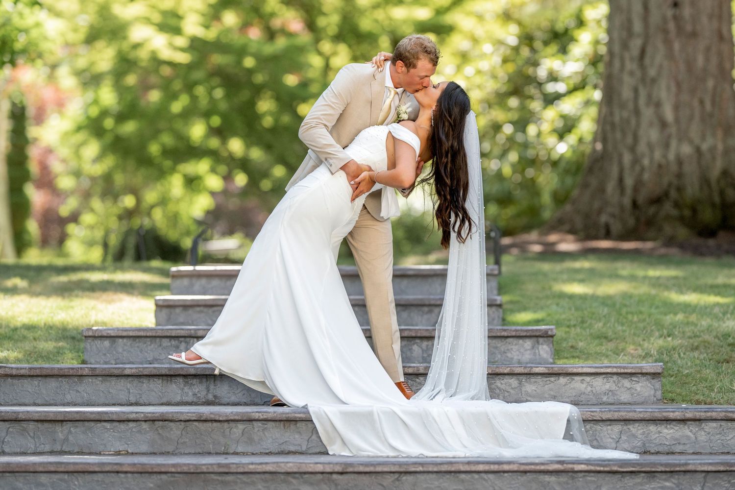 Bride and groom share a dip kiss on stone steps surrounded by lush green trees in outdoor wedding photo.