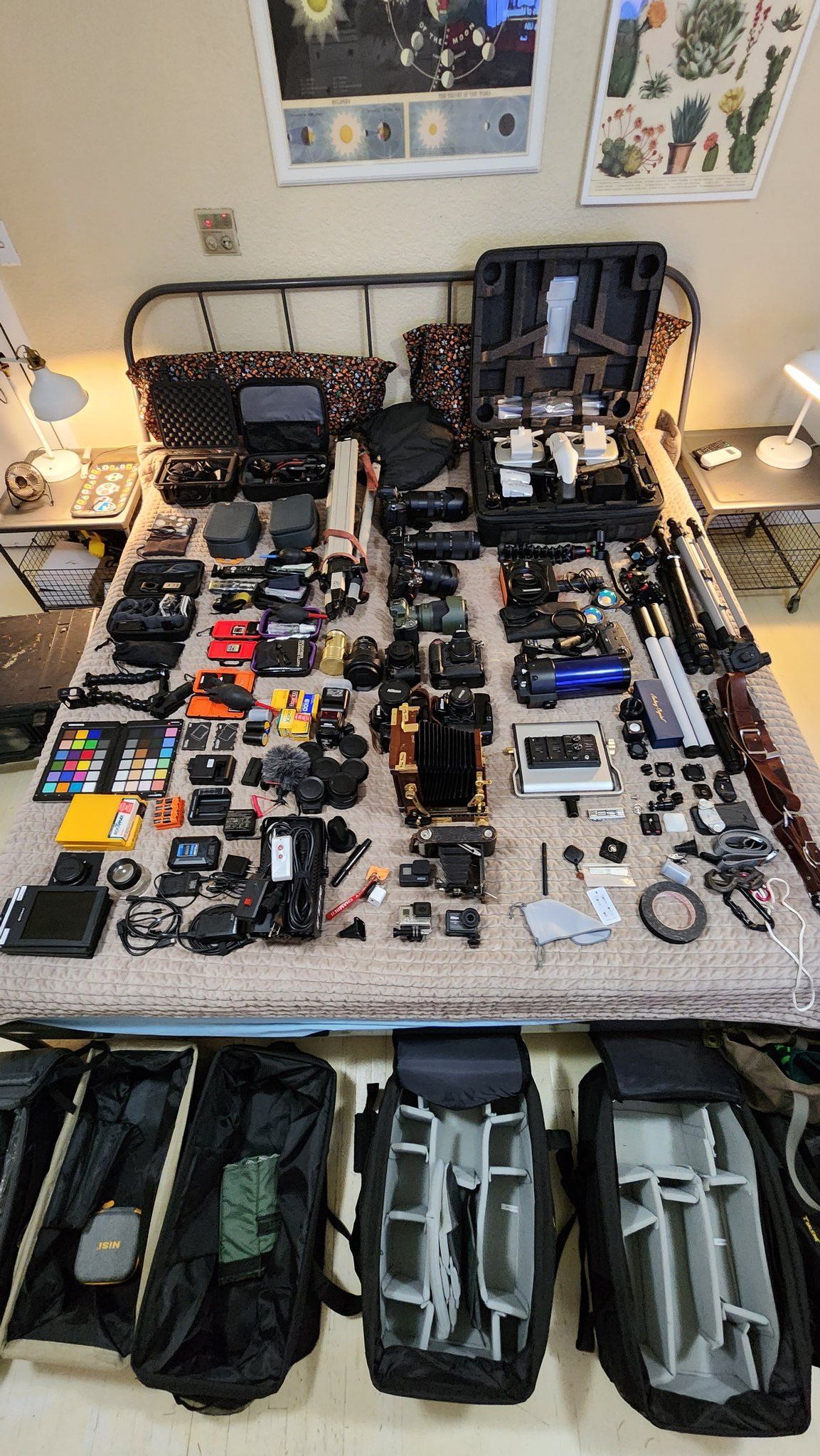 Professional camera gear collection laid out on a bed, including cameras, lenses, tripods, color charts, and carrying cases.