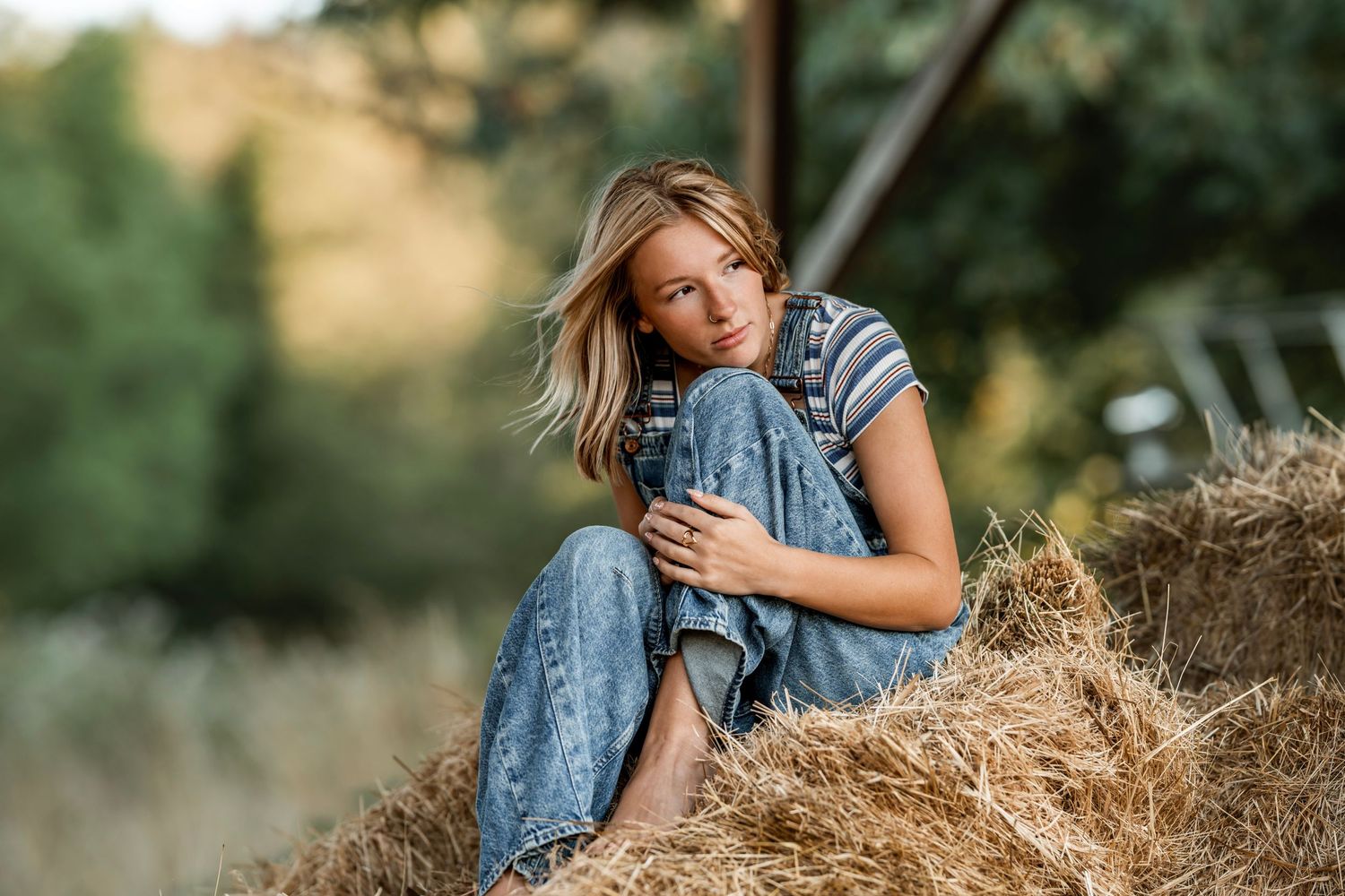 Young blonde woman sitting on hay bale in jeans and striped tee, rustic outdoor farm setting.