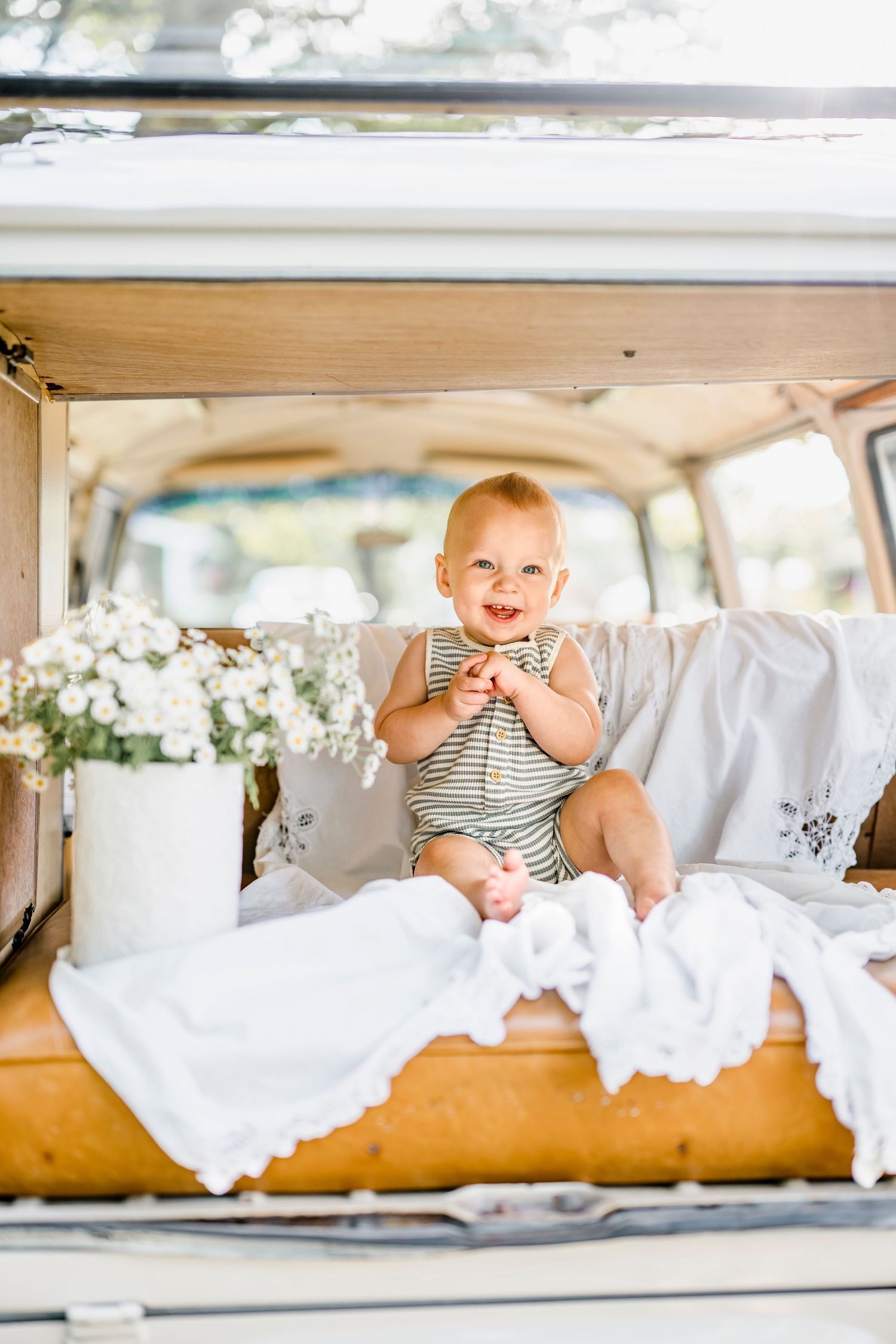 Happy baby in striped romper sitting in vintage van trunk with white flowers and blankets.