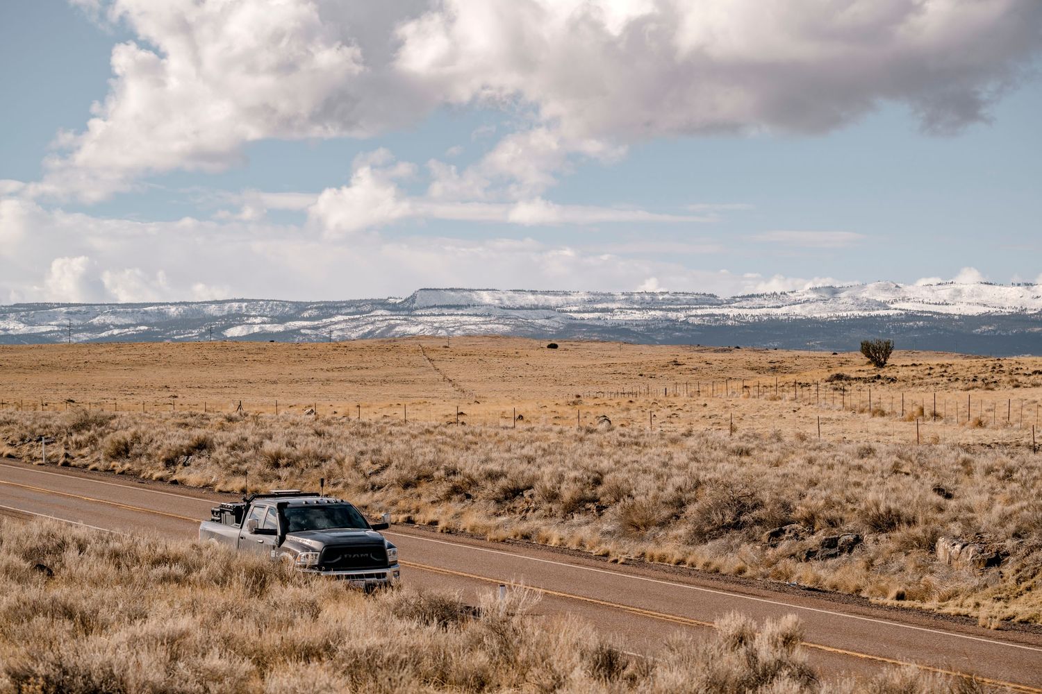 Silver pickup truck drives down rural highway through vast open plains with snow-capped mountains beyond.