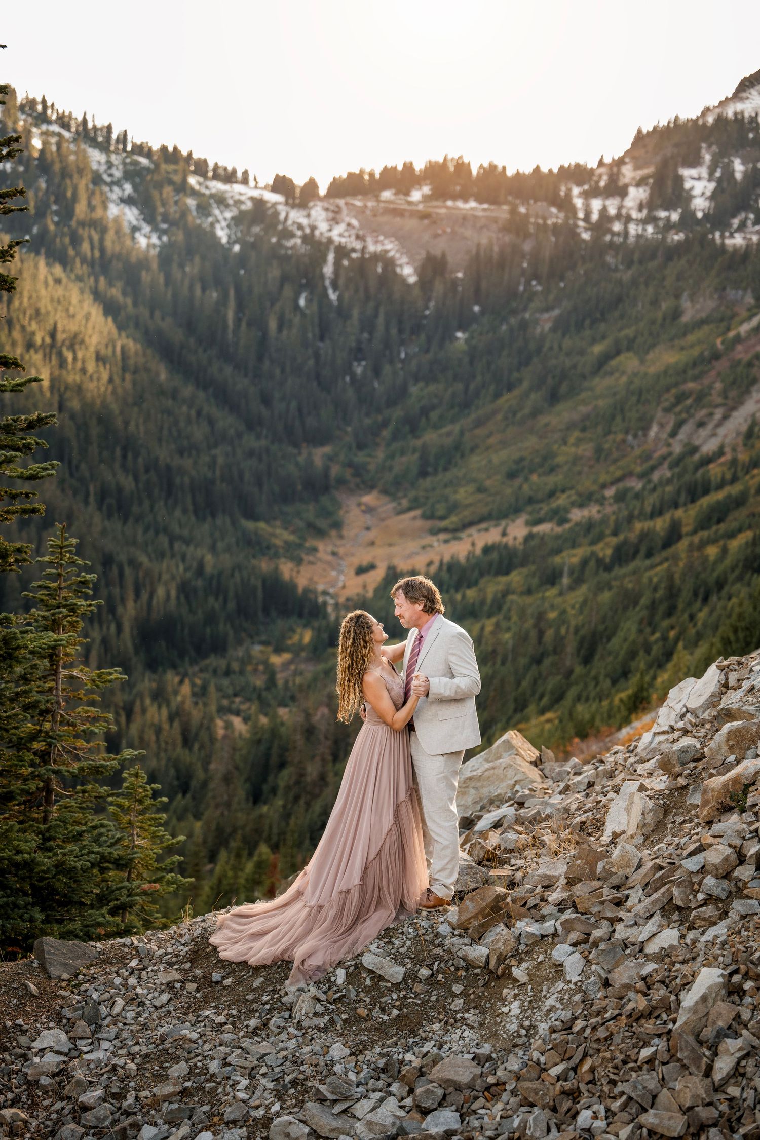 Couple in formal attire embracing on a rocky mountain slope with snow-capped peaks at sunset.