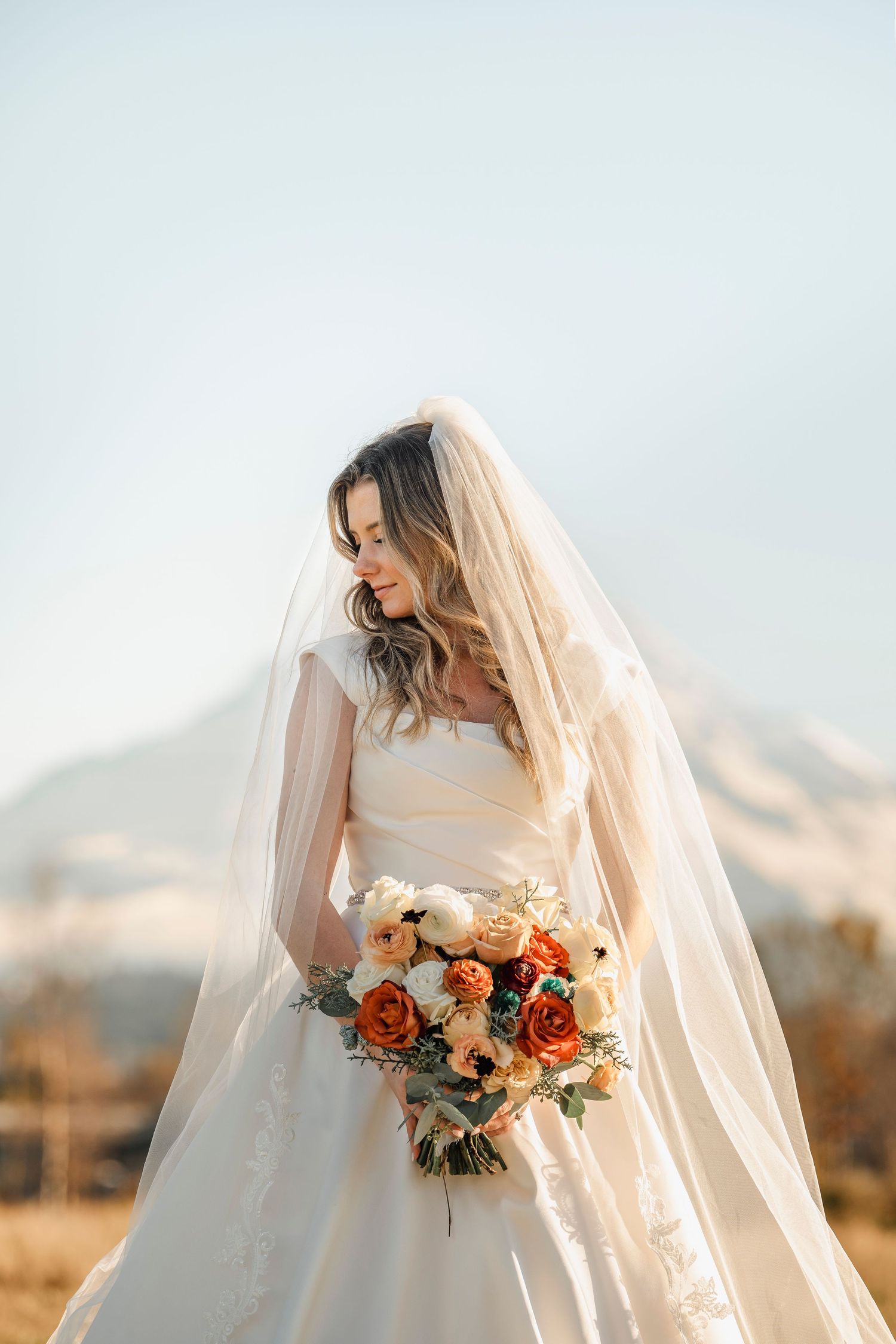 Bride in white gown and veil holds autumn bouquet of orange and cream roses with mountain backdrop.