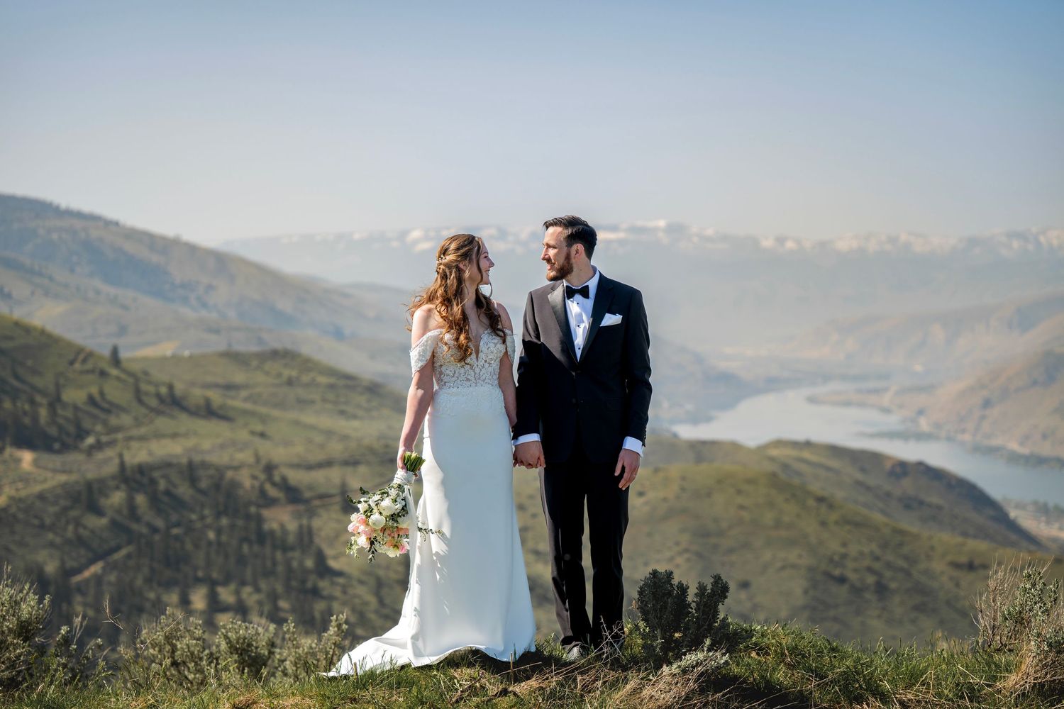 Bride and groom hold hands on mountain summit with scenic valley view.