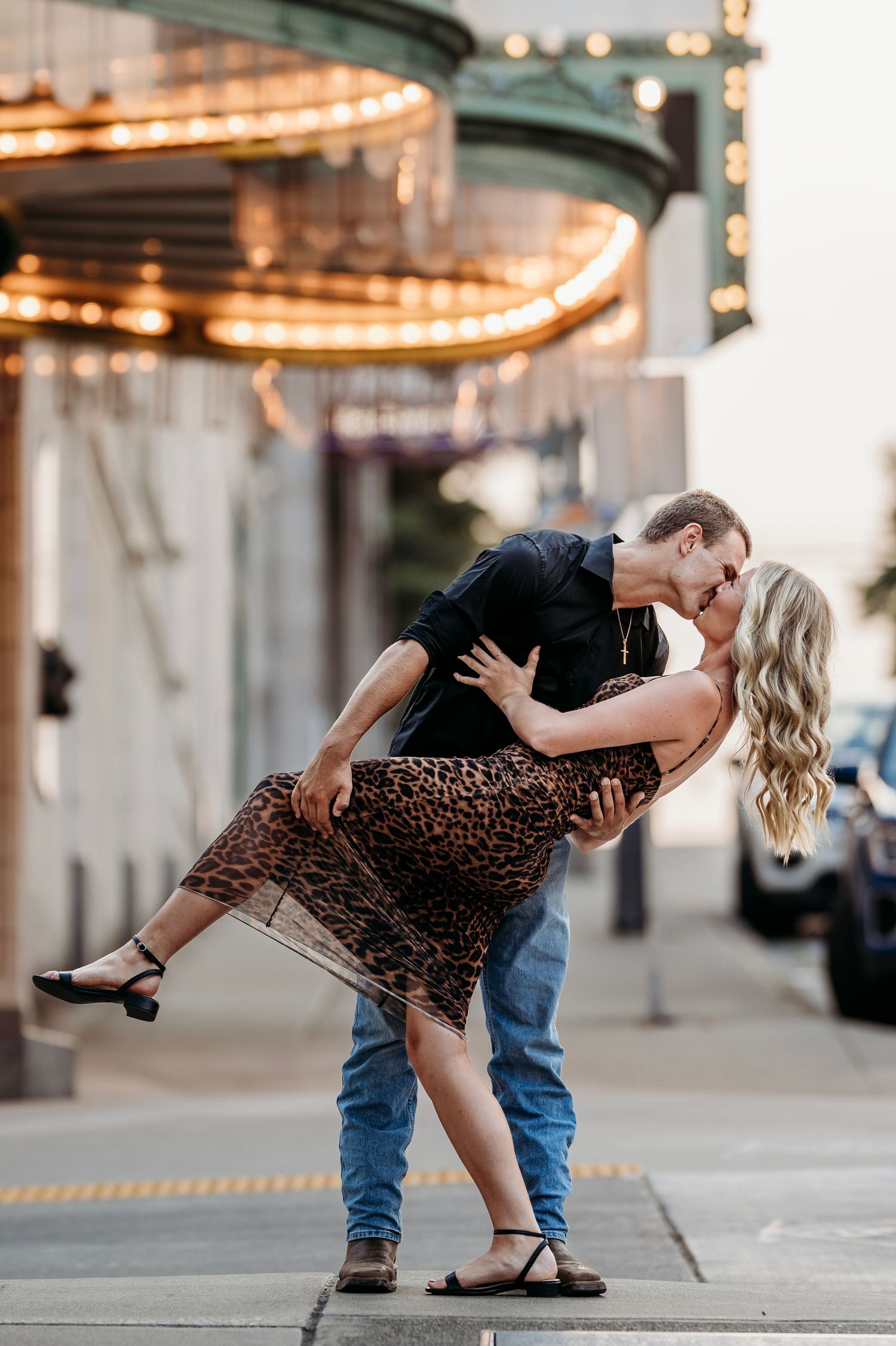 Couple sharing a romantic dip kiss on a city sidewalk under marquee lights at dusk.