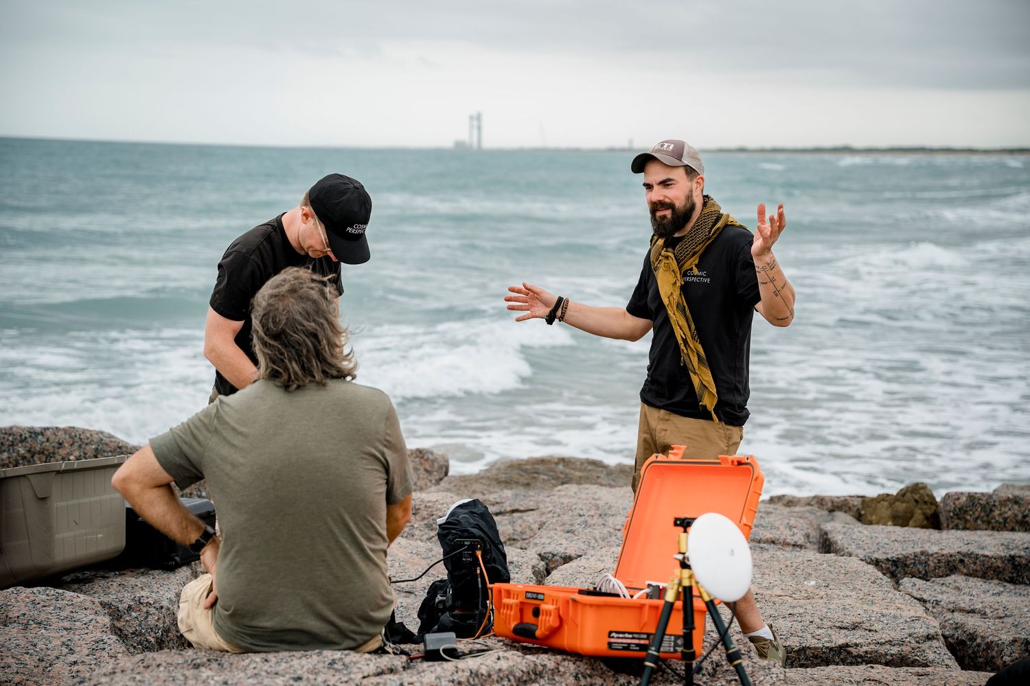 Bearded filmmaker gesturing while briefing crew on rocky ocean shoreline with orange equipment case.