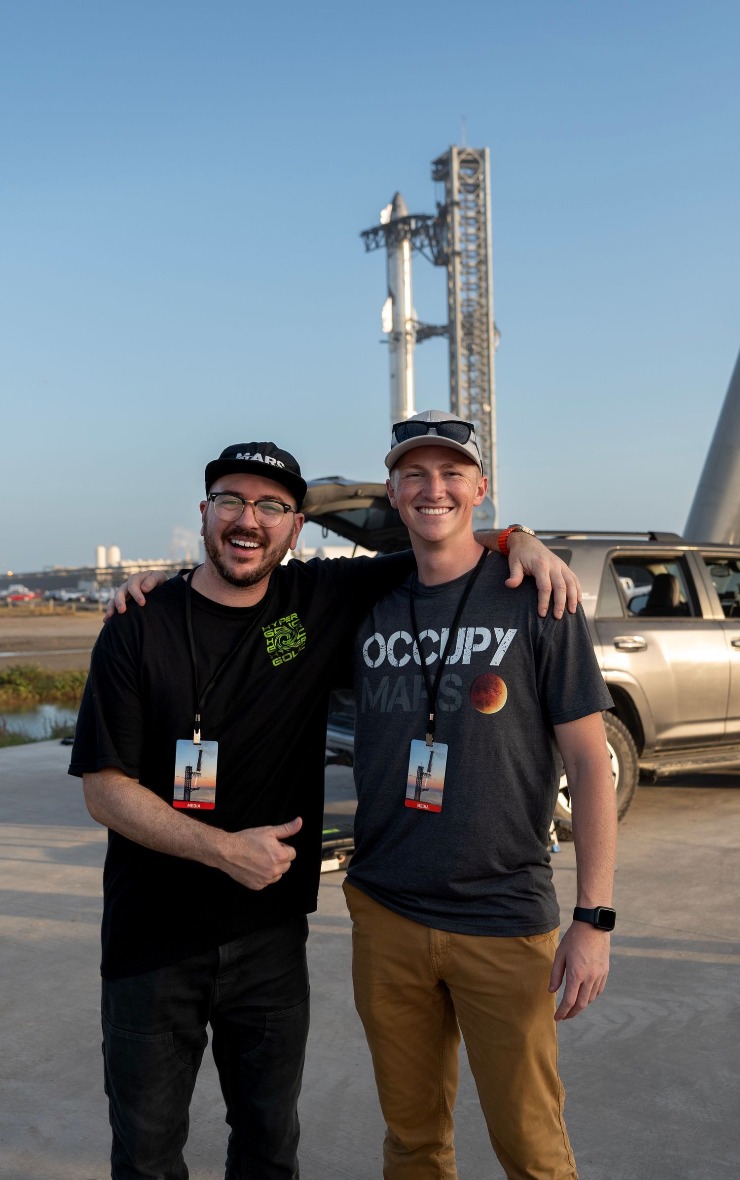 Two smiling men wearing Mars-themed shirts pose together in front of a SpaceX rocket launch facility.