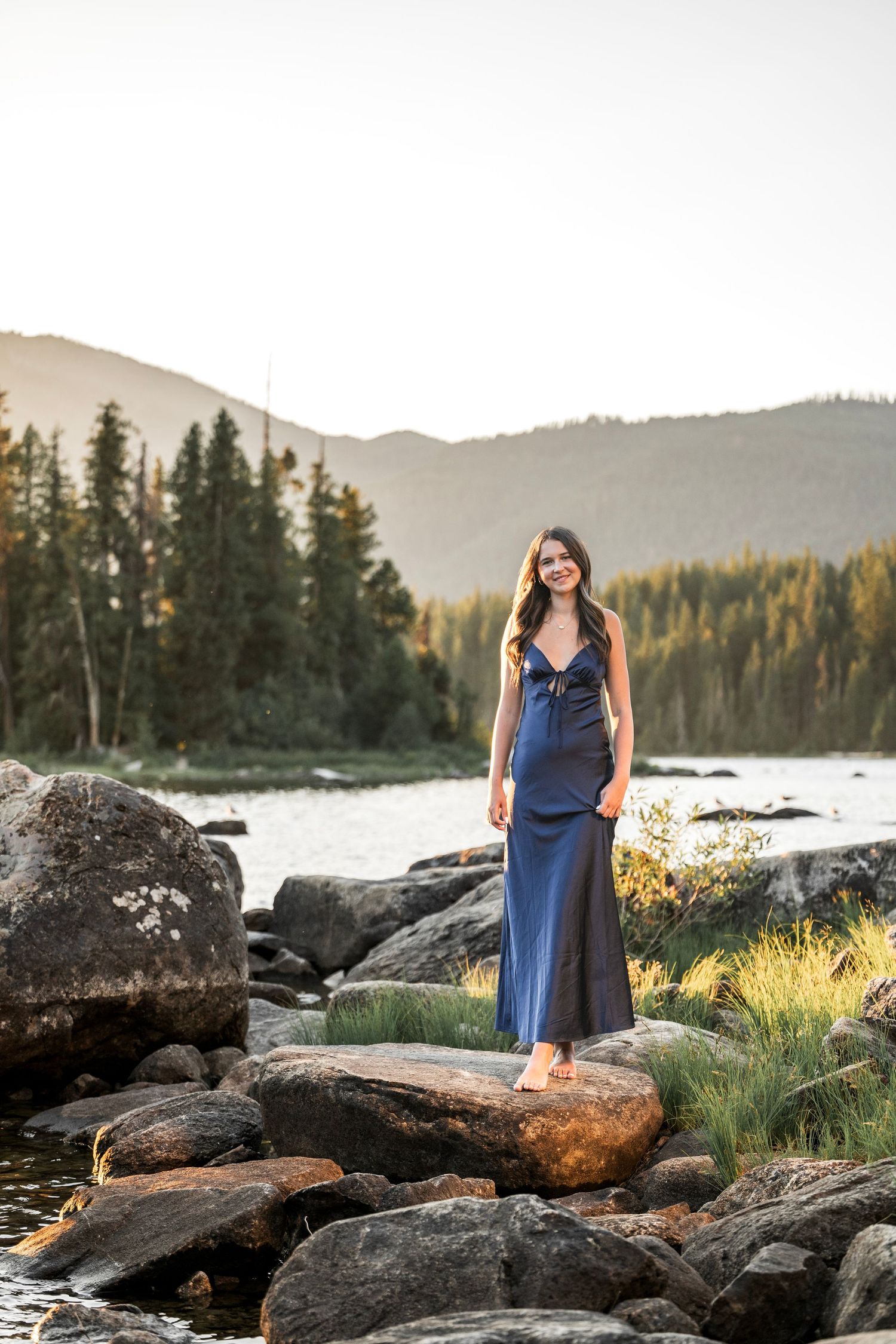 Young woman in navy blue maxi dress standing on rocks by a mountain lake at golden hour sunset.