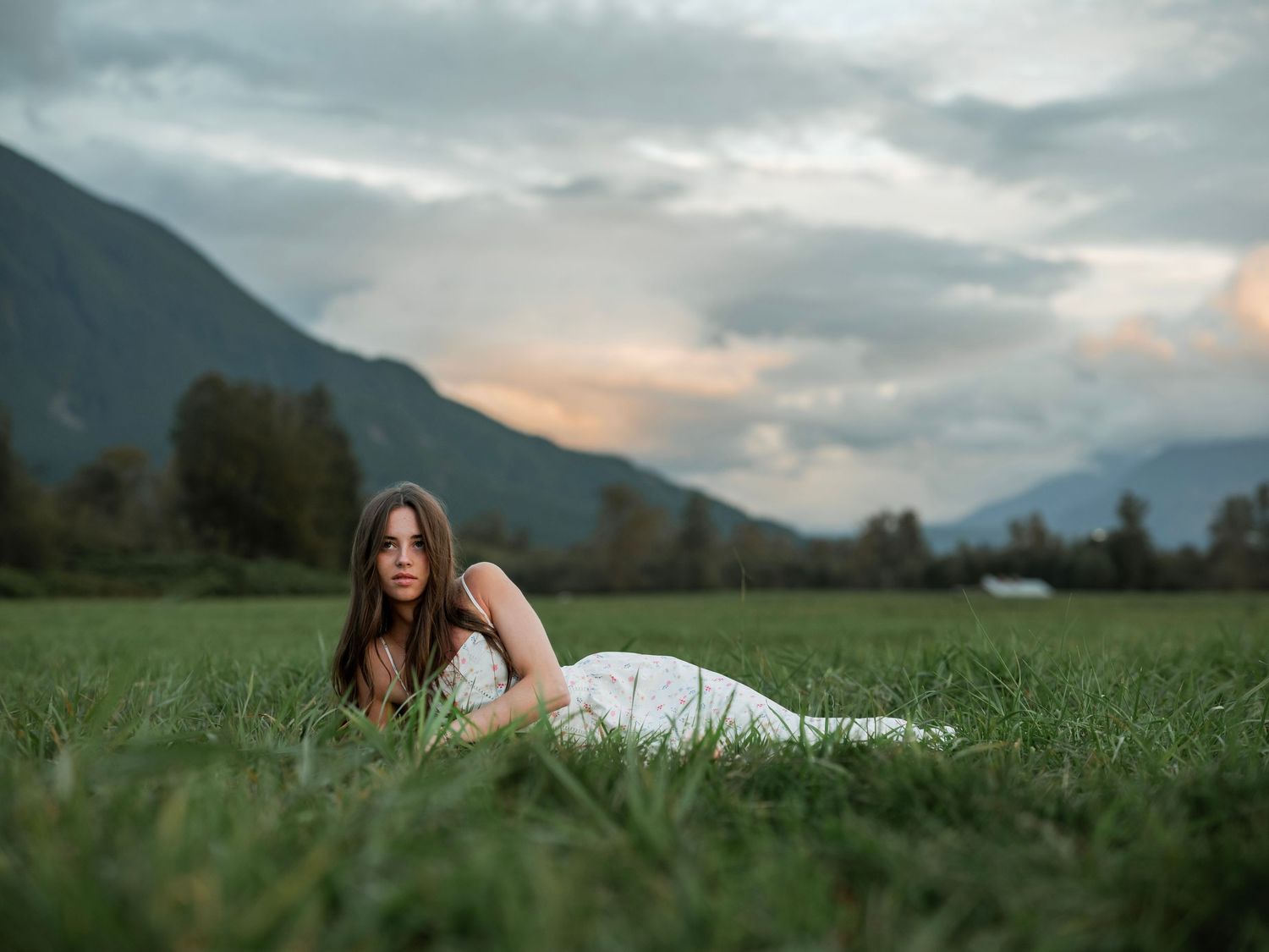 Young woman in white dress lying on lush green grass with mountain backdrop and dramatic cloudy sky at dusk.
