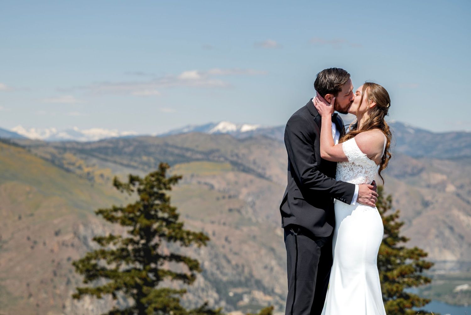 Bride and groom kissing on mountain summit with snow-capped peaks behind them.