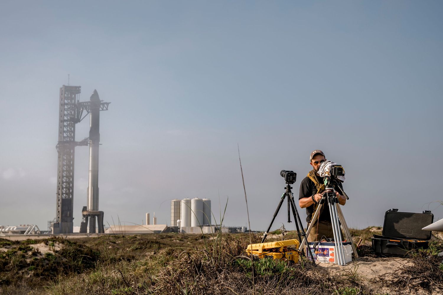 Photographer with camera on tripod near SpaceX Starship launch tower at Boca Chica, Texas on a hazy day.