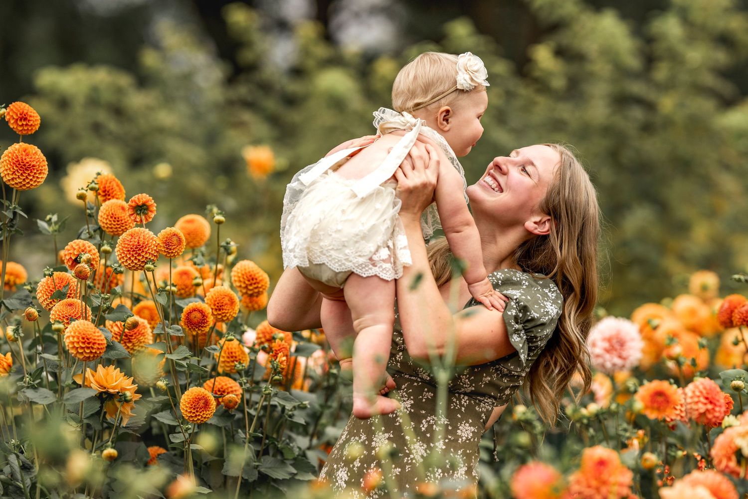 Mother joyfully lifting smiling baby girl among vibrant orange dahlia flowers in a lush garden setting.
