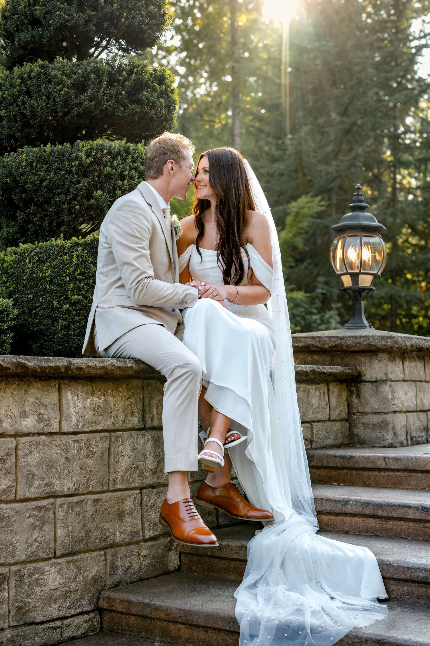 Bride and groom sharing a romantic moment on stone steps beside a lantern in a lush garden at sunset.