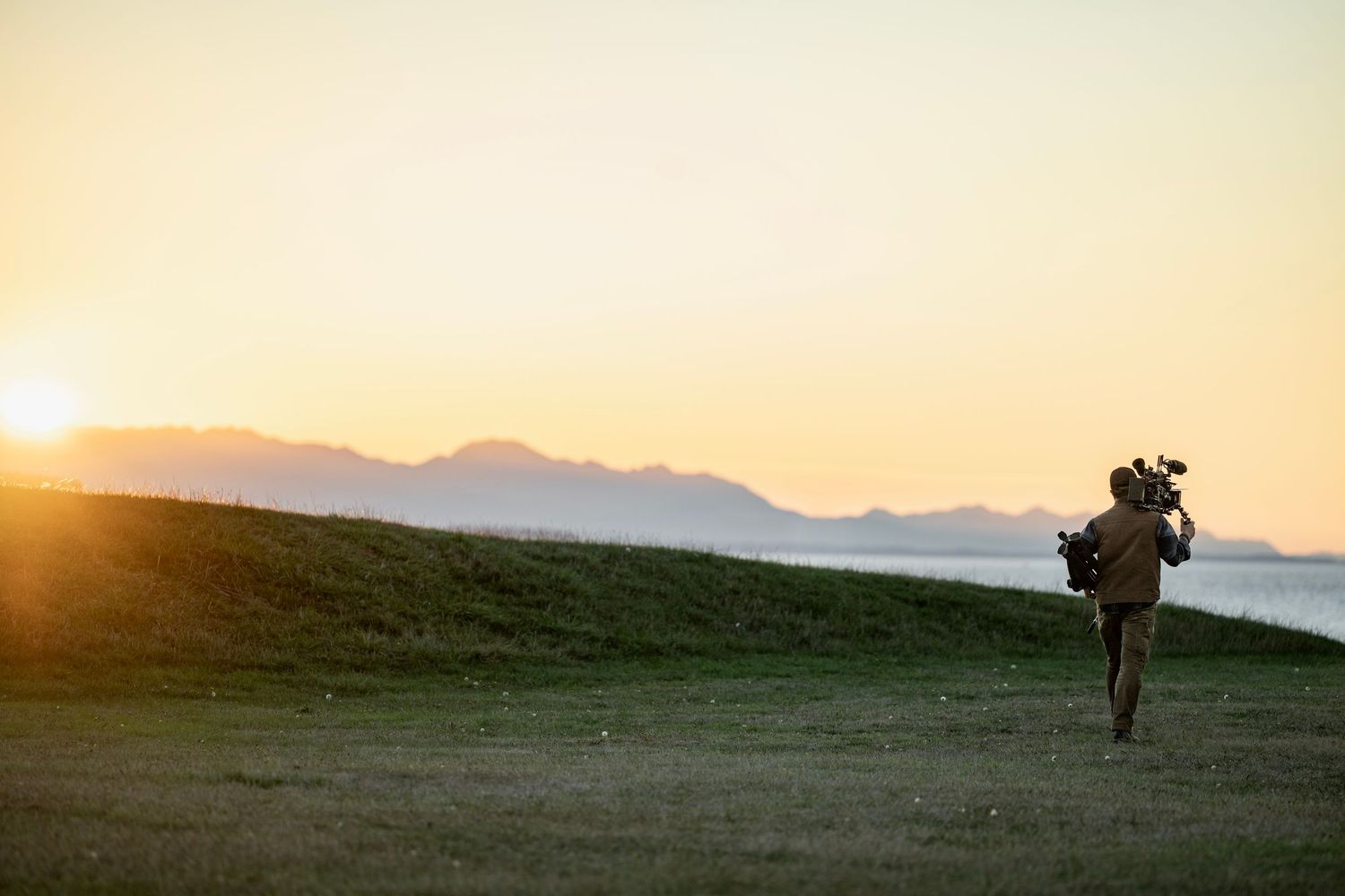 Silhouette of photographer carrying equipment walks across green golf course at golden sunset by the sea.
