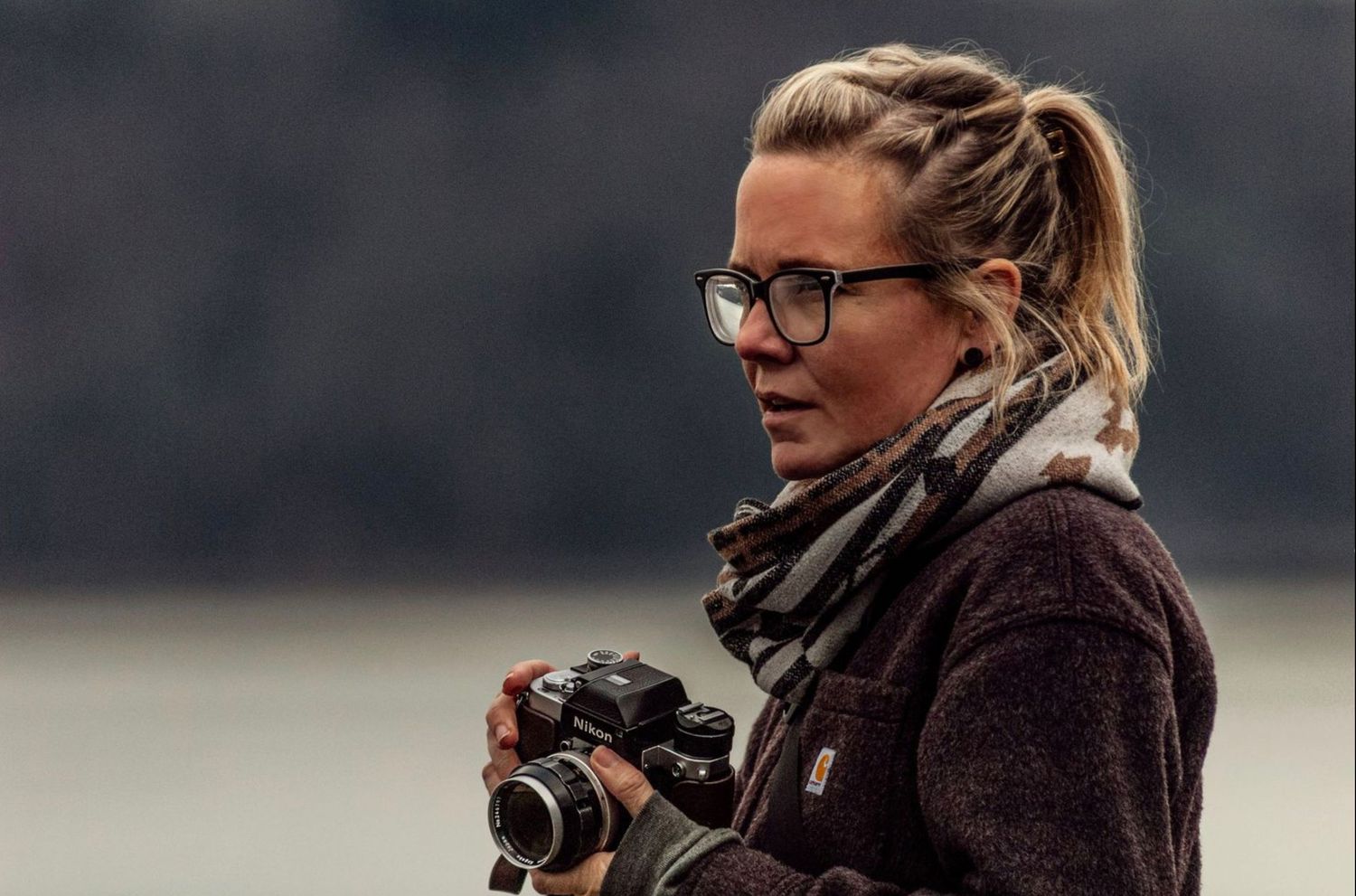 Focused female photographer with glasses and scarf holds camera outdoors near misty lake in autumn.