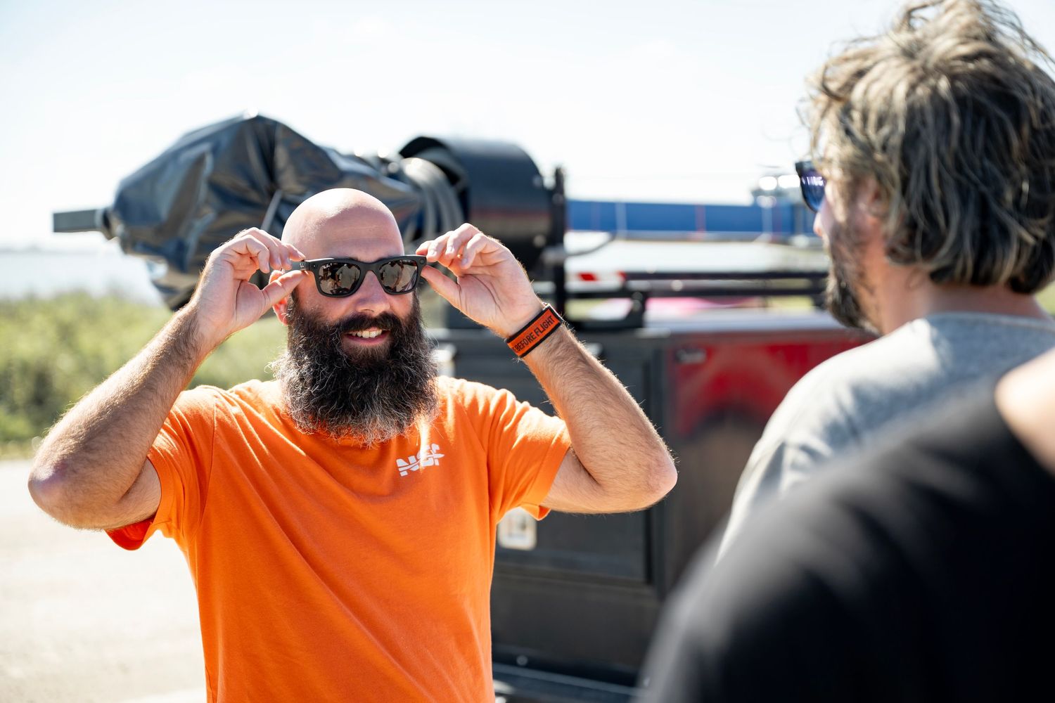 Bearded man in orange shirt tries on sunglasses outdoors, smiling near a vehicle in sunny weather.