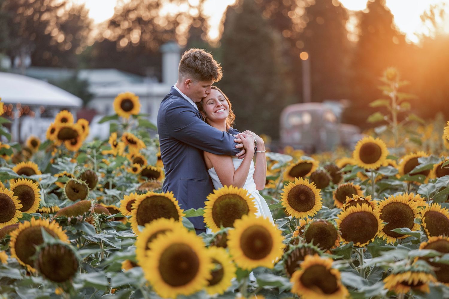 Couple embracing joyfully in a sunflower field at golden hour, groom in blue suit hugging bride in white dress.