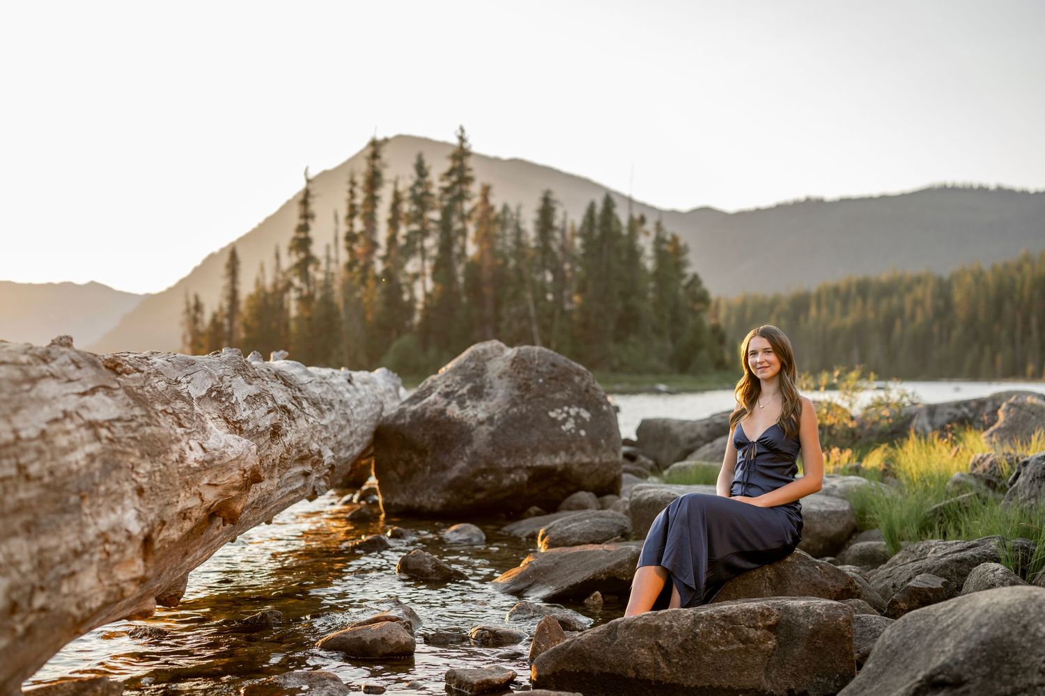 Young woman in a navy dress sitting on rocks by a mountain stream at sunset.