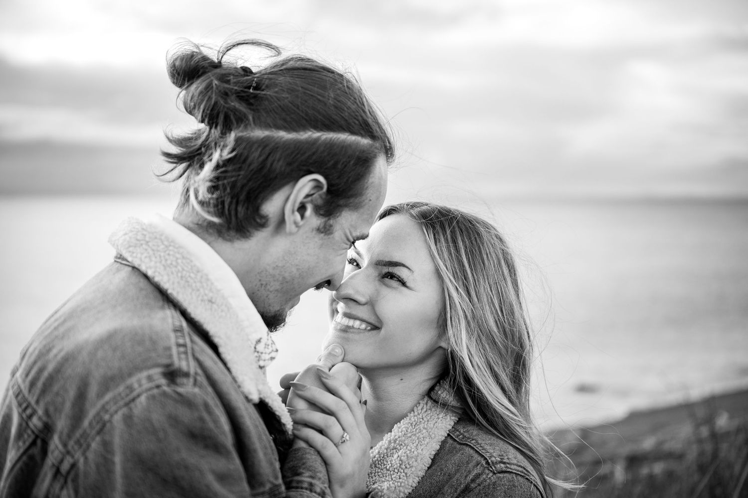 A romantic black and white photo of a couple nearly kissing with the ocean and cloudy sky in the background.