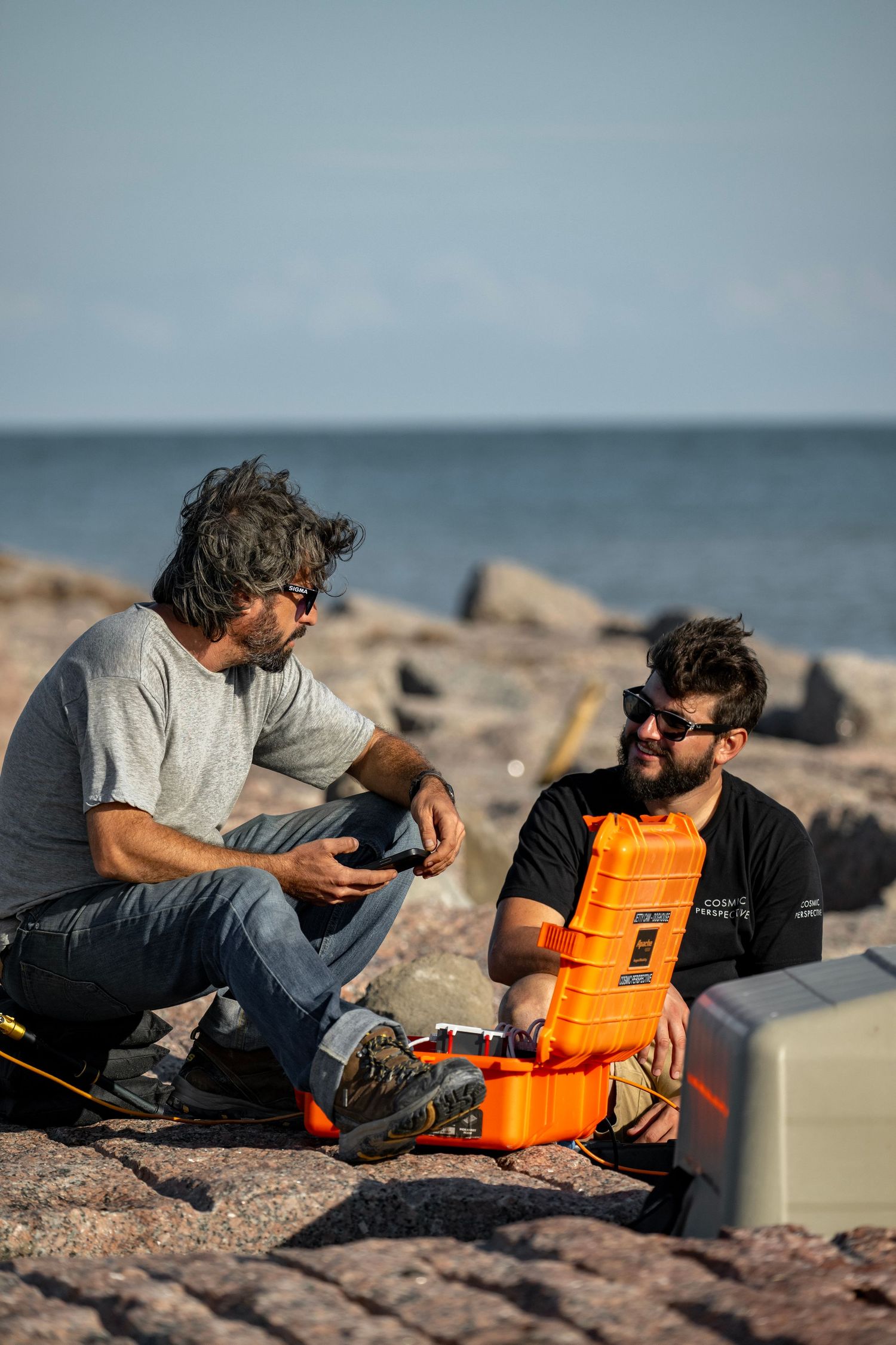 Two men sit on rocky shoreline conversing beside an orange equipment case with the ocean in the background.