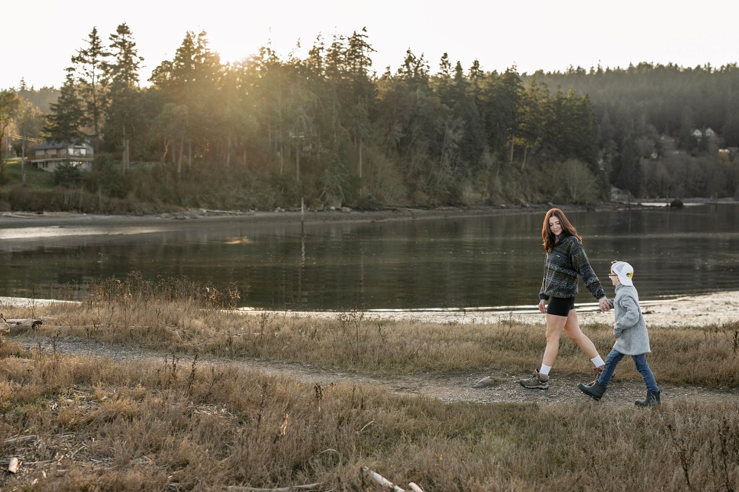 A mother and child walk hand in hand along a scenic lakeside path at golden hour with trees in background.