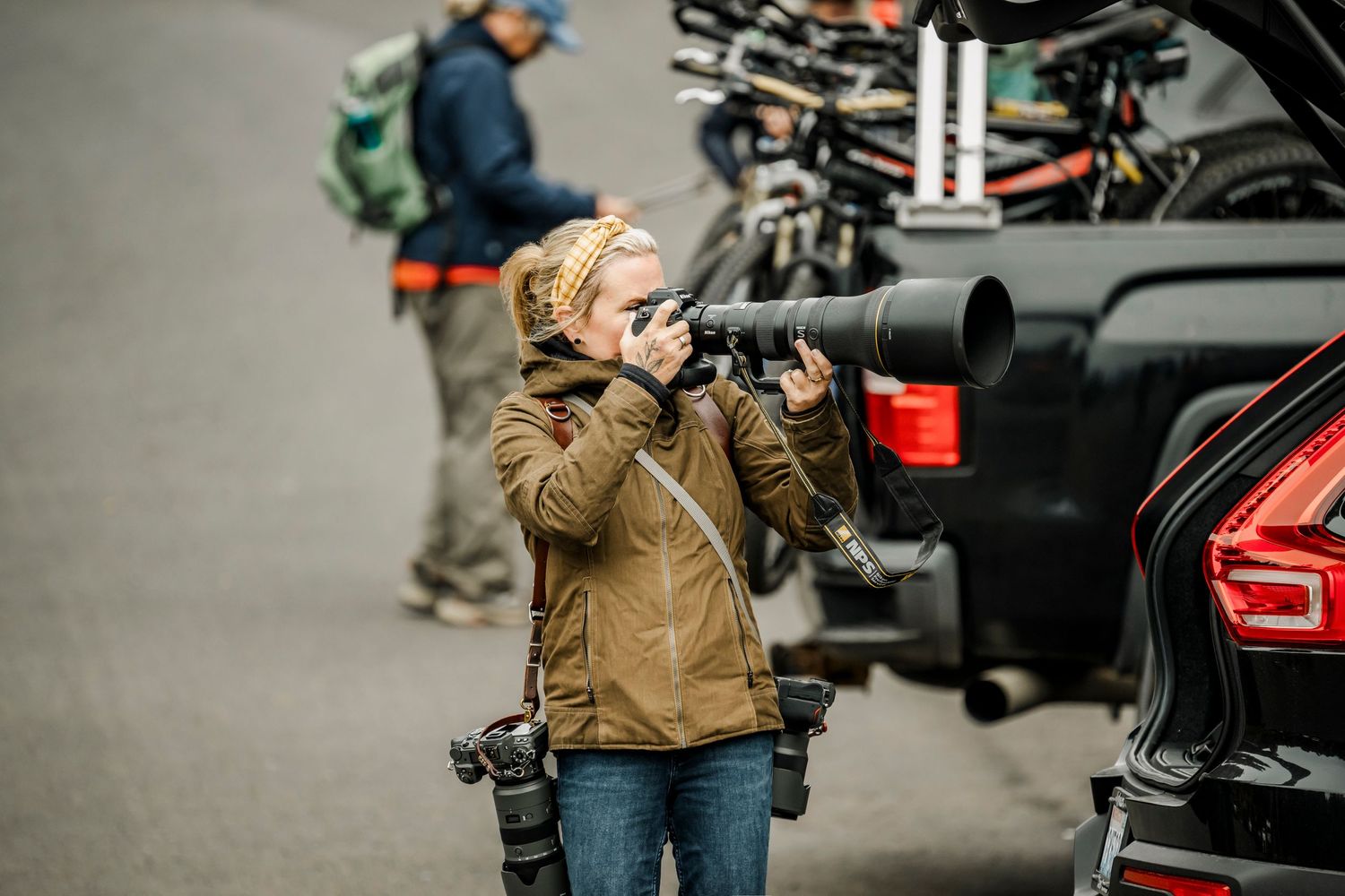 Female photographer with telephoto lens and camera gear shooting outdoors near vehicles at an event.