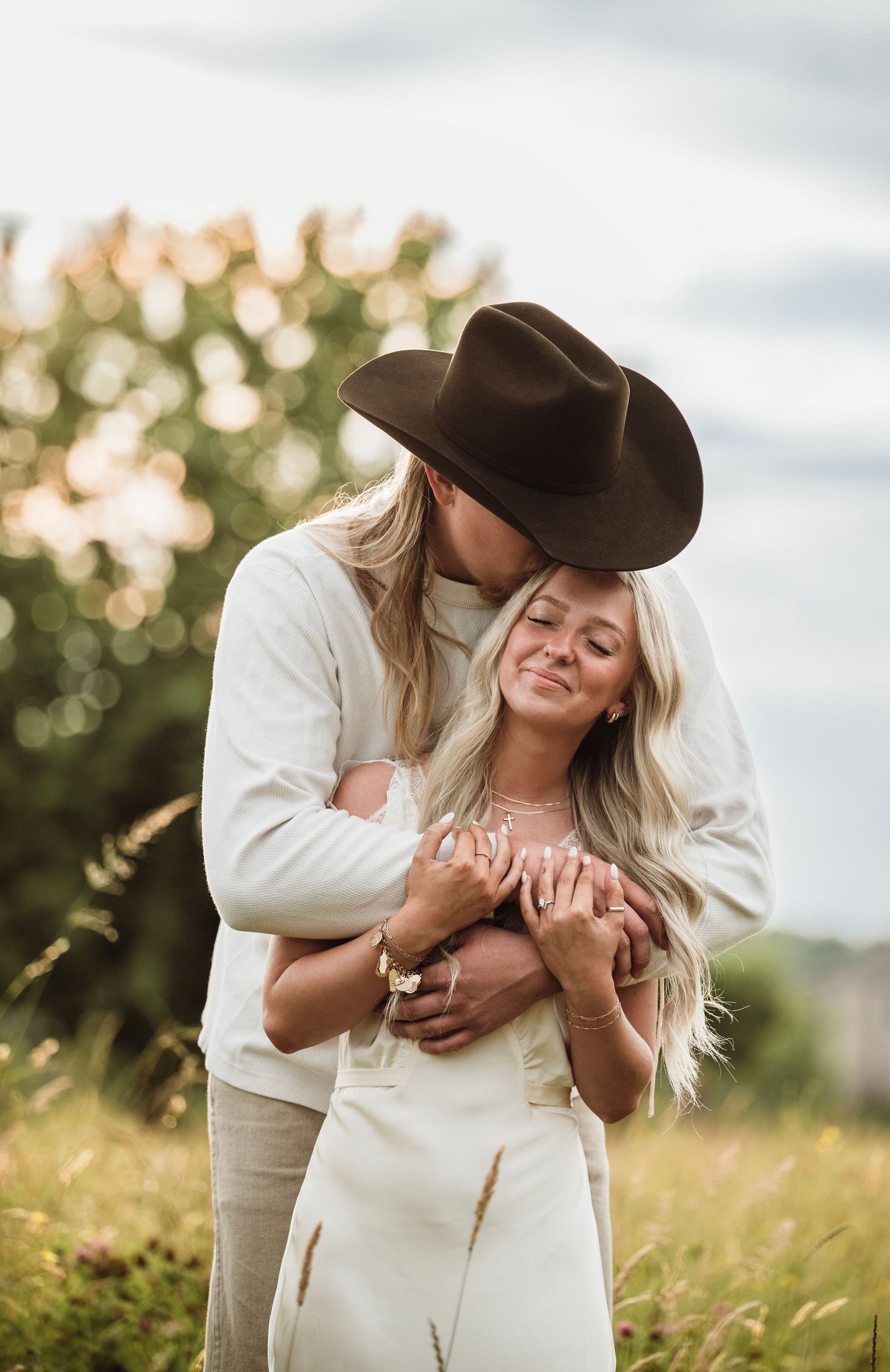 Couple embracing in a field, man in cowboy hat kissing woman's head while holding her close.