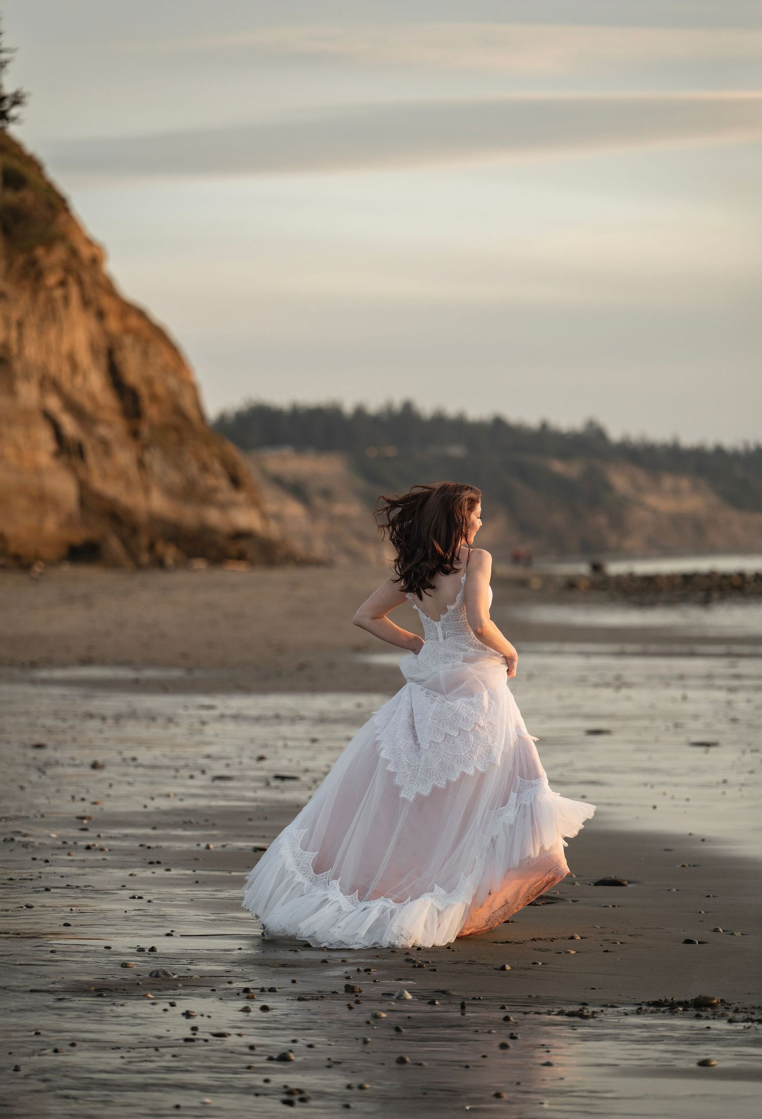 Bride in a flowing white gown walking along a scenic beach at golden hour sunset.