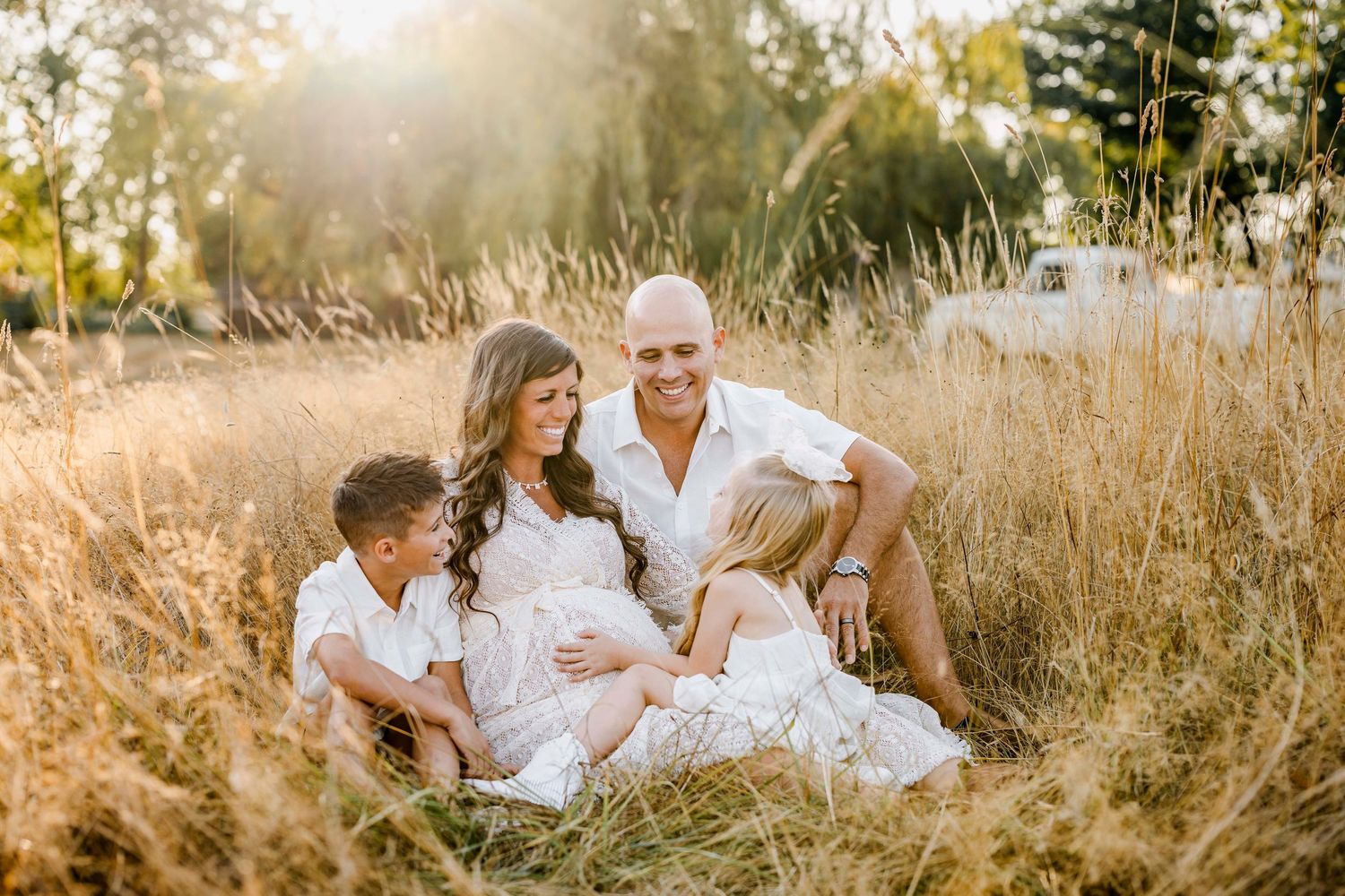 Family of four sitting in golden field, parents with two children in white outfits, warm sunlight glowing through tall grass.
