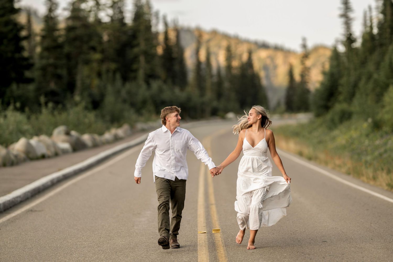Couple holding hands walking down a mountain road, woman in flowing white dress, man in white shirt.